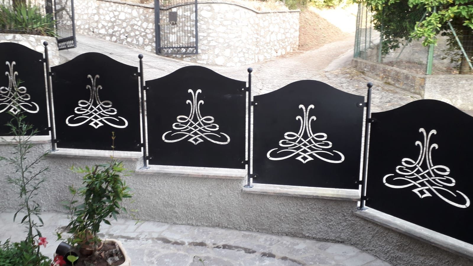Recinzione in ferro battuto nero con ritagli decorativi bianchi, di fronte a un muro di pietra e a un sentiero in pendenza.
