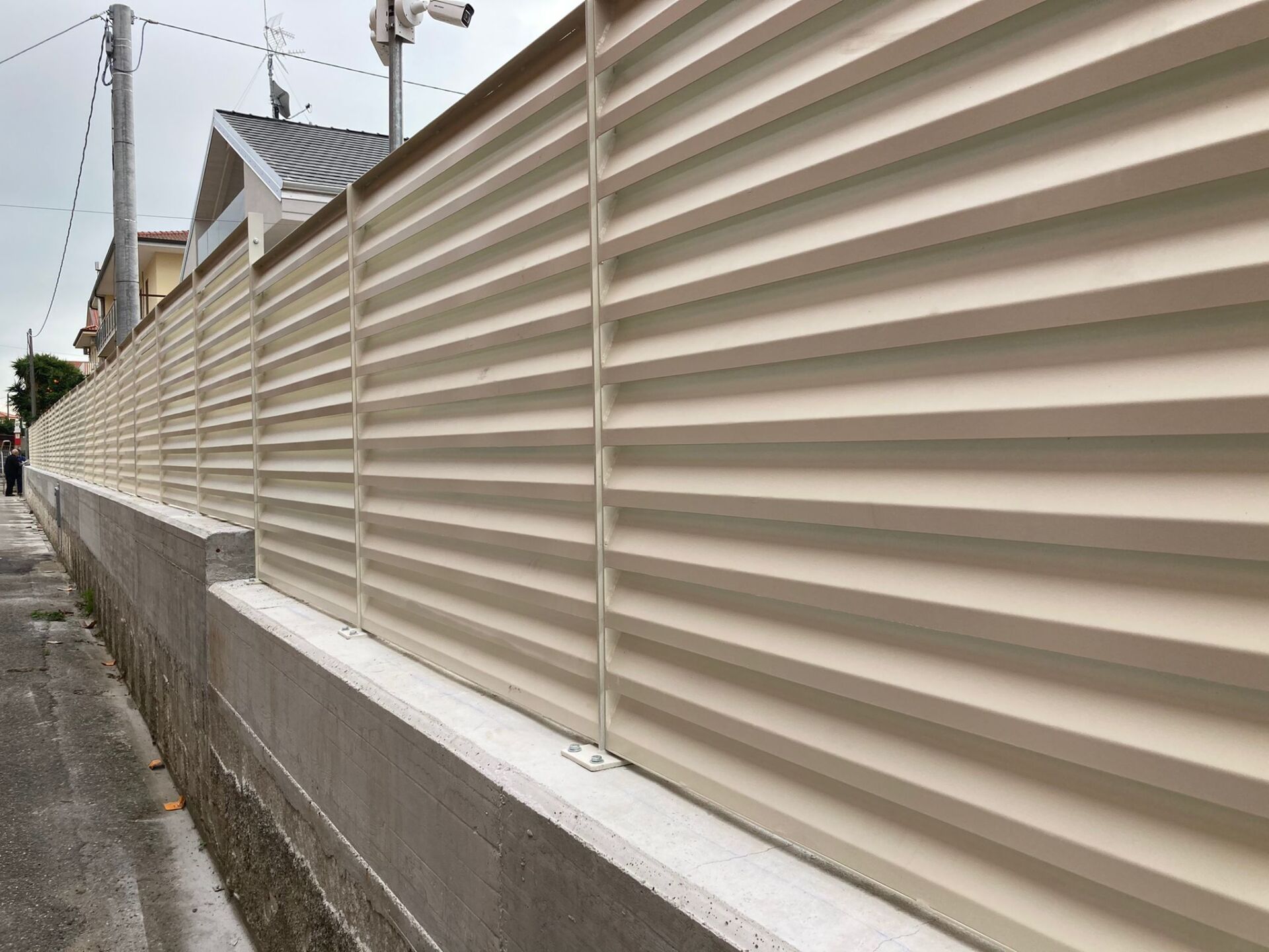 Recinzione orizzontale beige a doghe in cima a un muro di cemento lungo un marciapiede, con una casa visibile sullo sfondo.