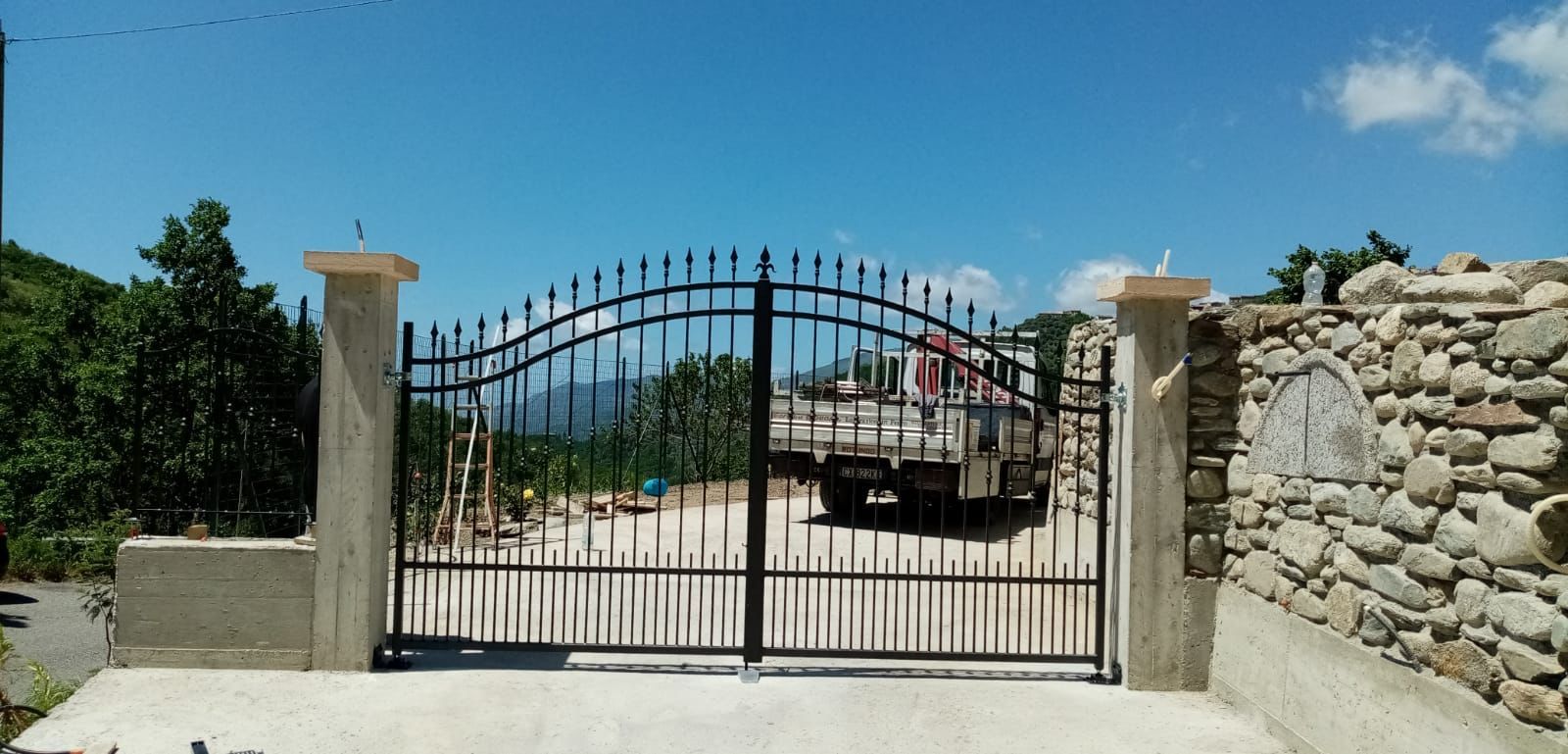 Cancello di metallo nero tra pilastri di pietra, che conduce a una zona montuosa sotto un cielo azzurro.