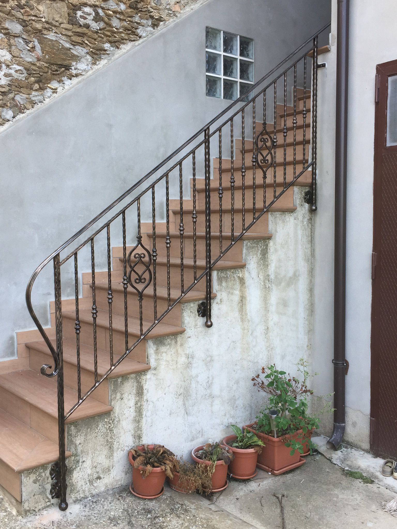 Scala con ringhiera in ferro battuto all'esterno di un edificio; gradini in terracotta, varie piante in vaso lungo la base.