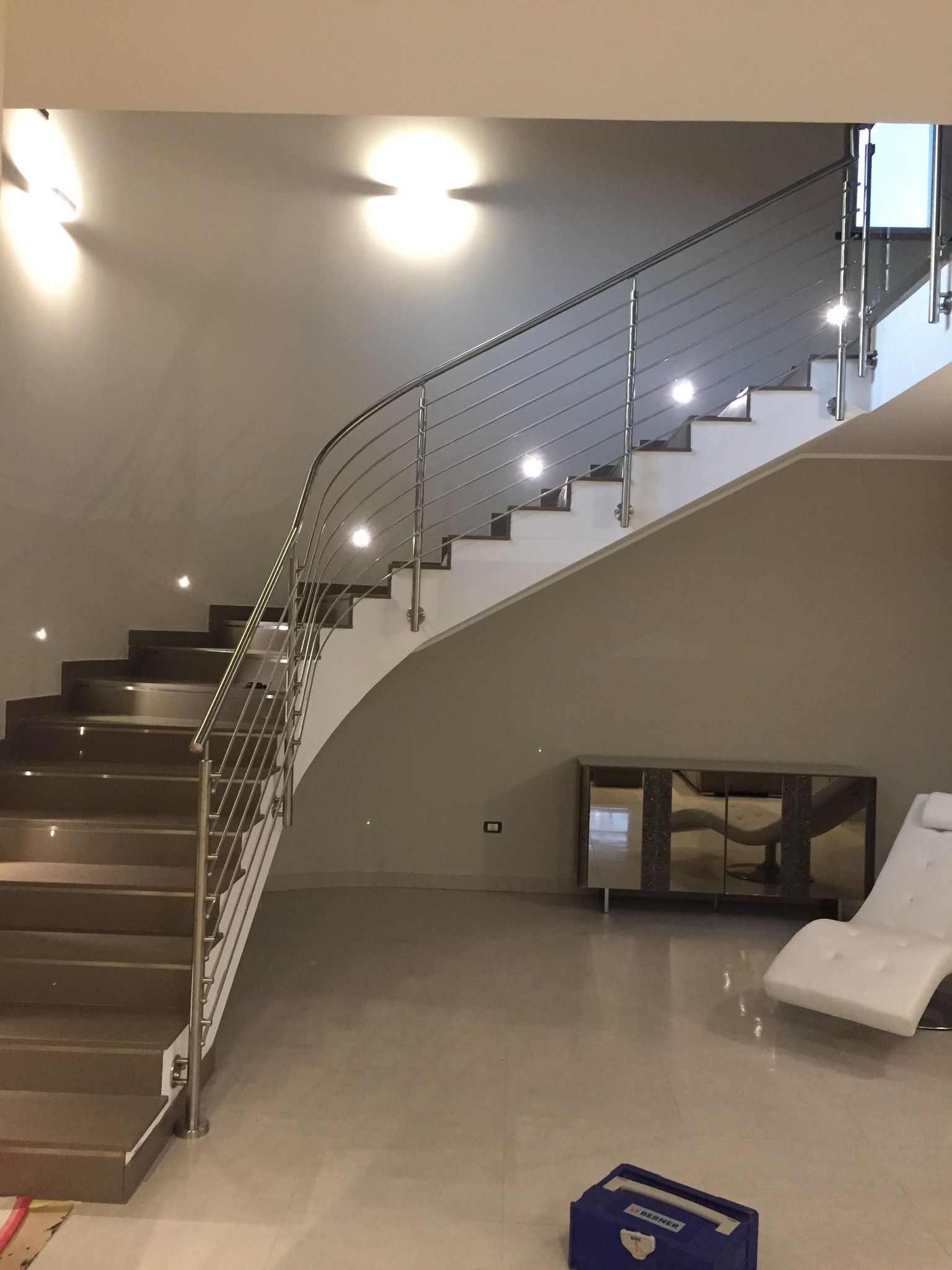 Moderna scala con ringhiera in acciaio inox e illuminazione integrata che conduce al piano superiore, pareti beige e pavimento piastrellato.