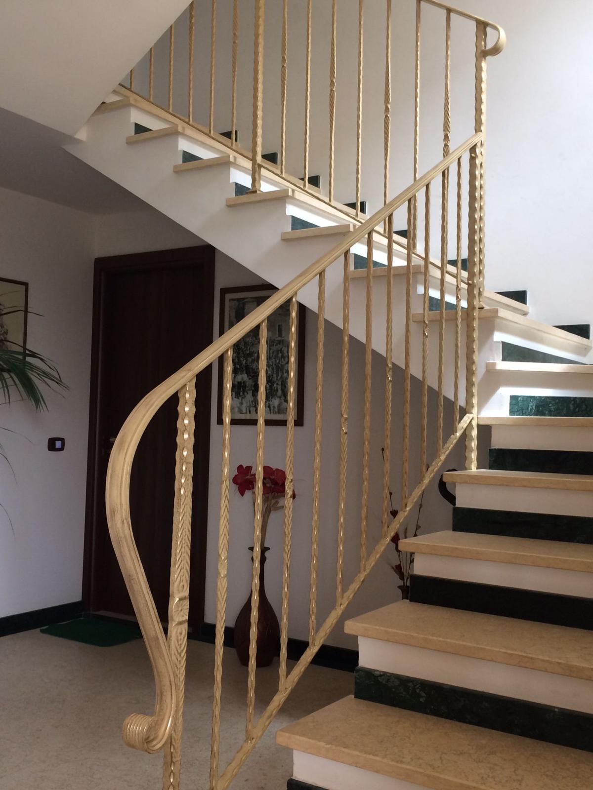 Una scala con ringhiere dorate. Gradini bianchi con bordi verdi conducono in alto, vicino a una porta di legno scuro.