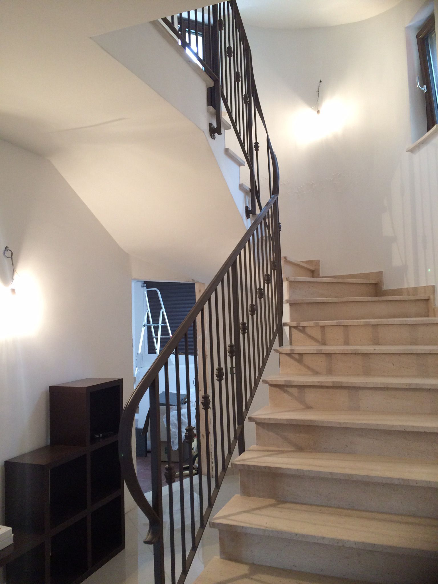 Scala con gradini in pietra color cuoio, ringhiera in ferro battuto, pareti bianche e scaffalature in legno scuro.