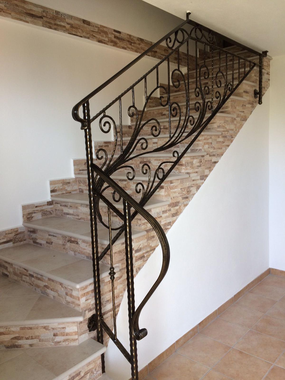 Scala con gradini in pietra marrone e ringhiera in ferro battuto nero decorato contro pareti bianche.