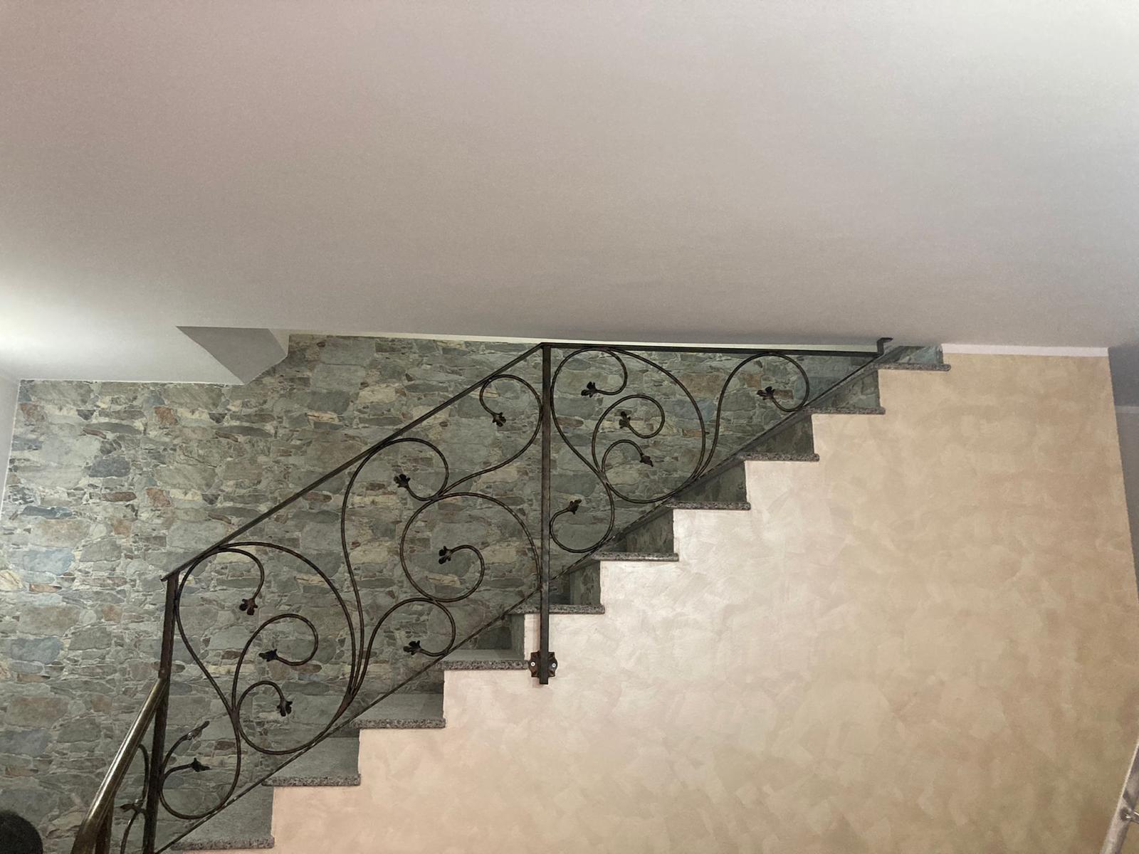 Scala con ringhiera in ferro battuto nero decorato. I gradini sono di colore chiaro con pareti ruvide su entrambi i lati.