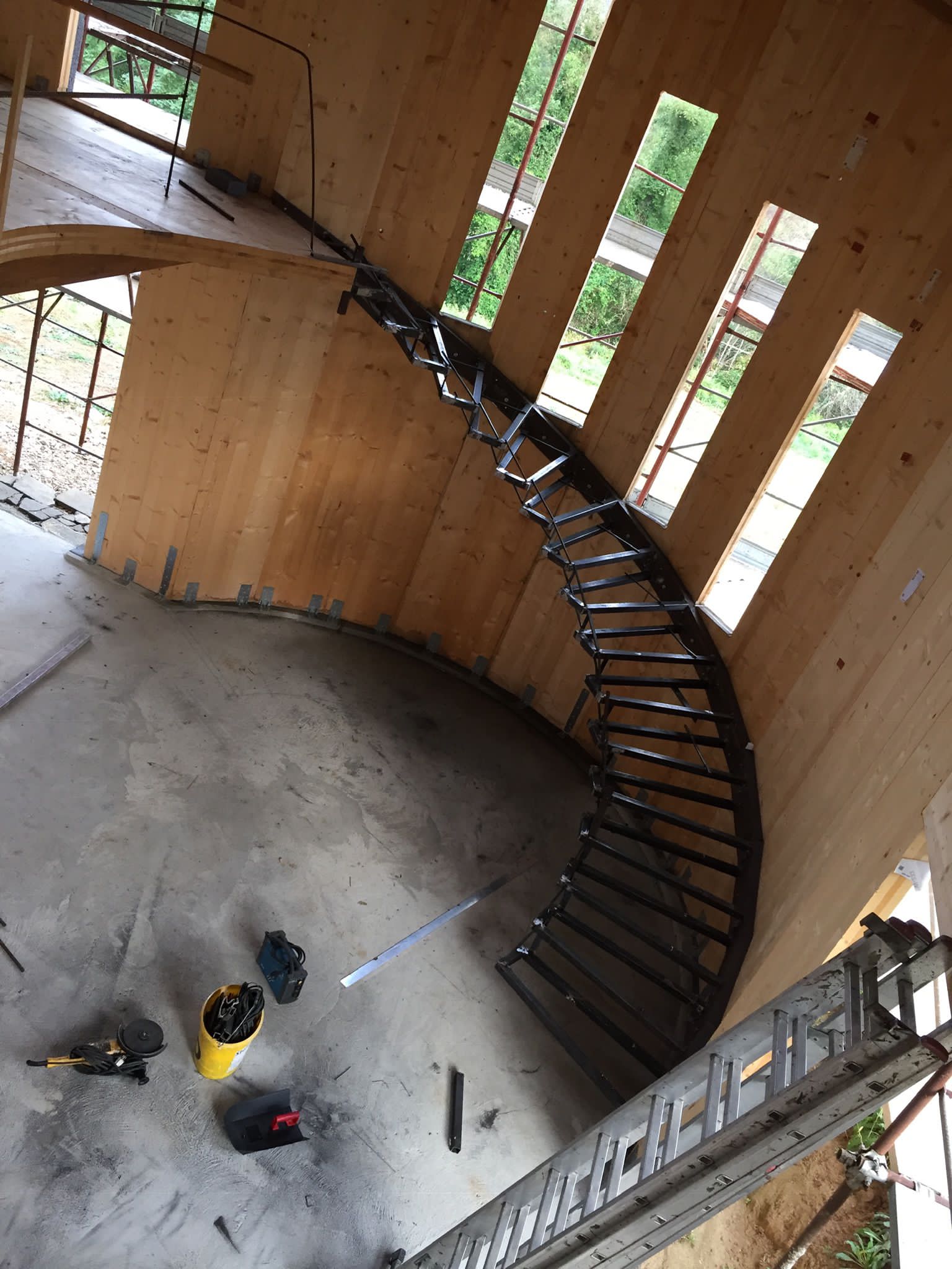 Scala curva in costruzione in una struttura con pareti in legno, con attrezzi visibili sul pavimento in cemento.