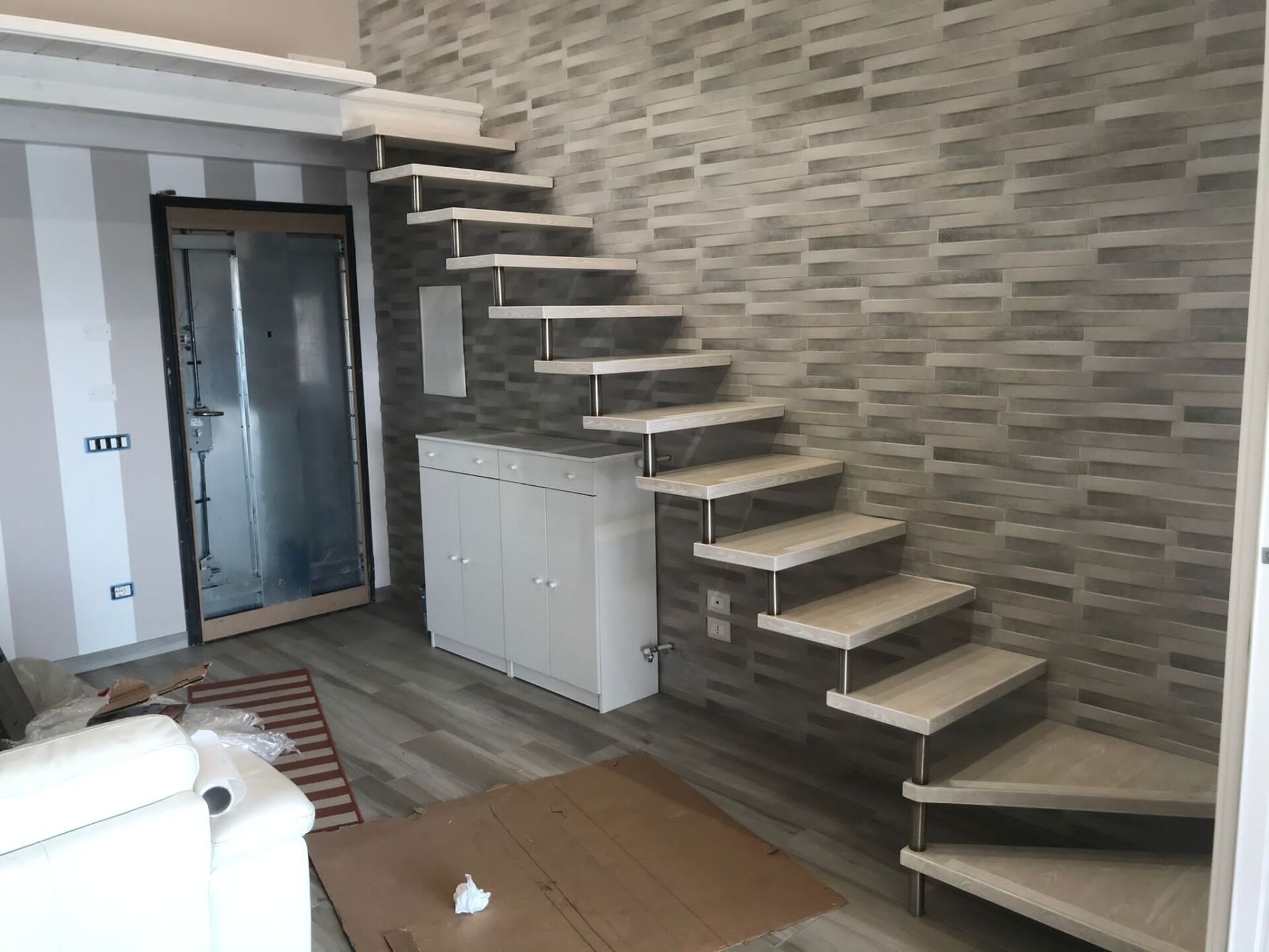 Scala sospesa con gradini chiari e supporti in metallo, contro un muro di mattoni grigi.