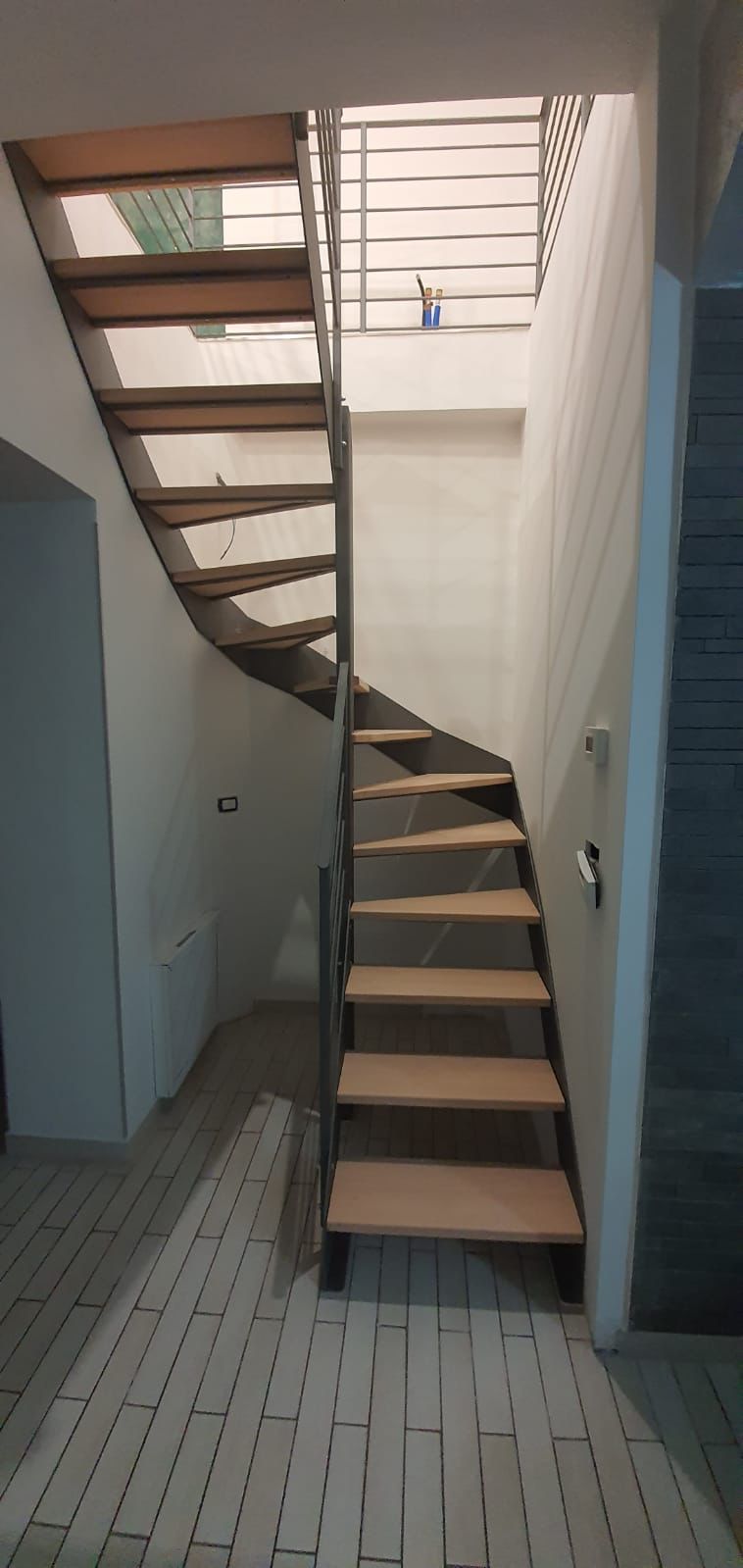 Una scala in legno con ringhiera in metallo conduce al piano superiore. Le pareti sono grezze. Il pavimento è piastrellato.