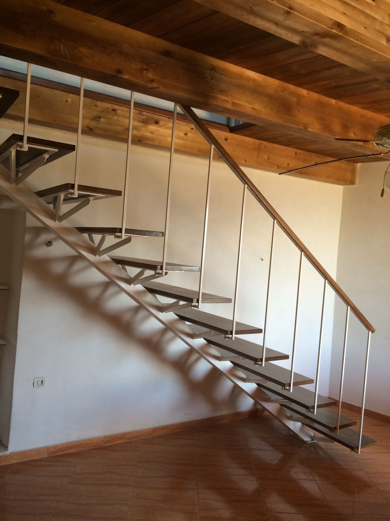 Scala in legno con supporti in metallo e corrimano contro una parete bianca e soffitto in legno.