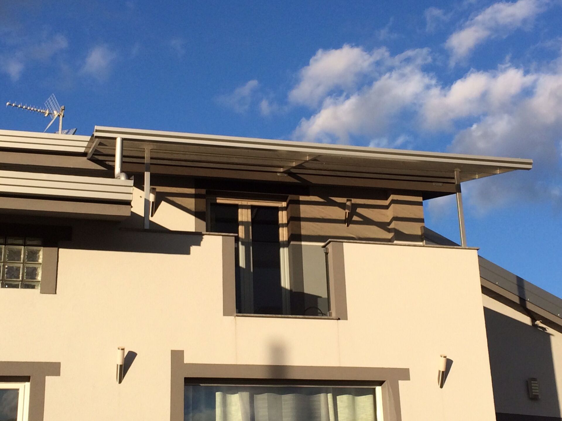 Esterno di una casa con tetto piano sopra una finestra, vernice grigia e bianca, cielo azzurro.