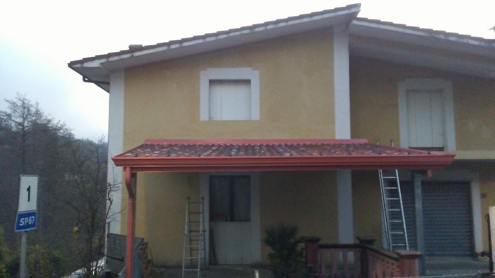 Edificio giallo a due piani con tetto in tegole rosse e portico. Due scale appoggiate alla struttura.