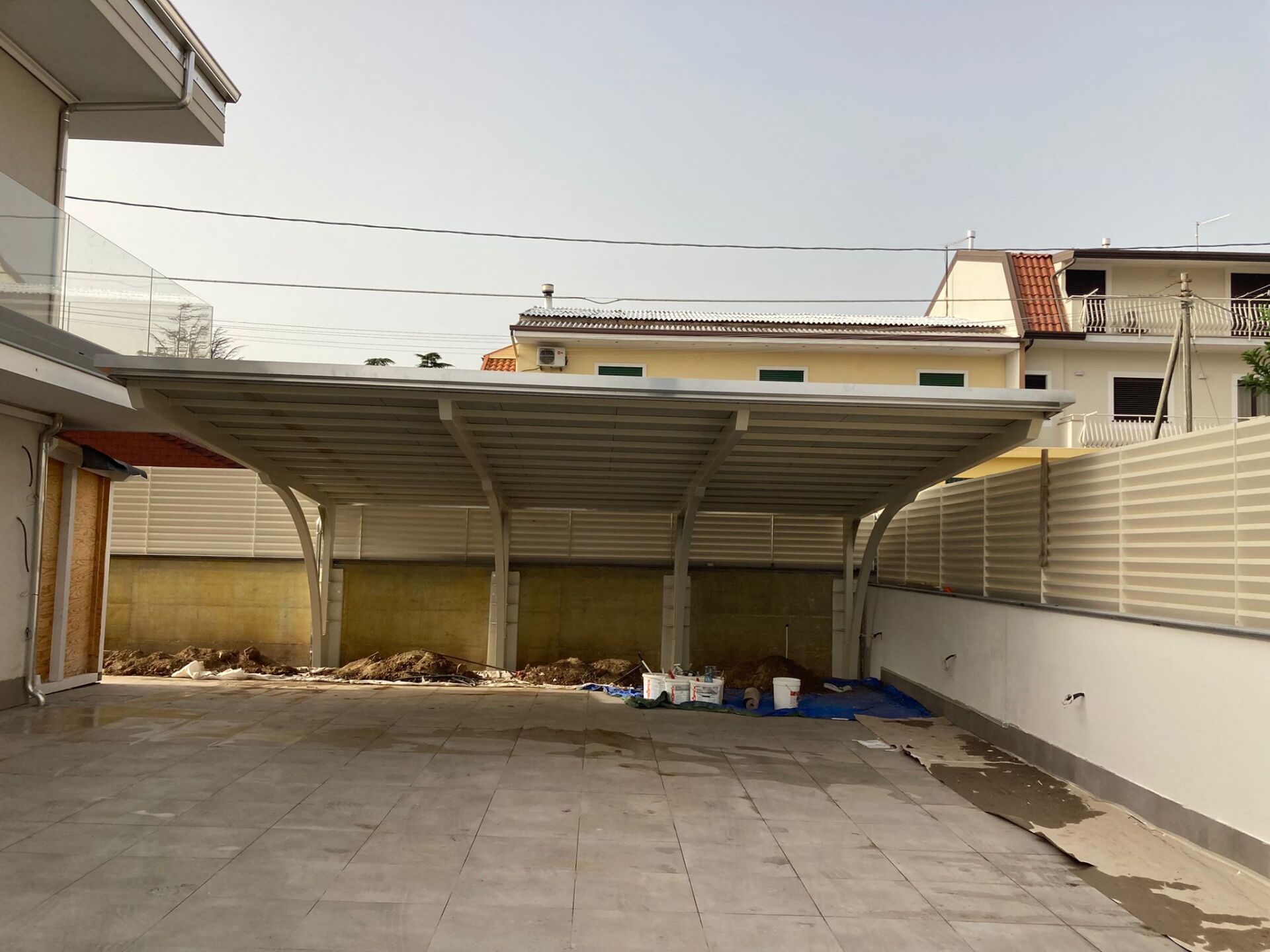 Carport con struttura in metallo e tetto chiaro, che protegge un'area pavimentata. Contesto residenziale con muri di cinta.