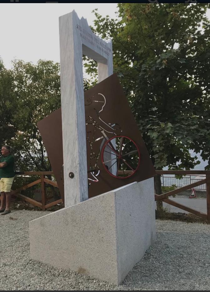 Monumento a un ciclista. Una figura ritagliata in metallo su una superficie inclinata, posta su una base di pietra, vicino ad alberi; accanto ad essa c'è un uomo.