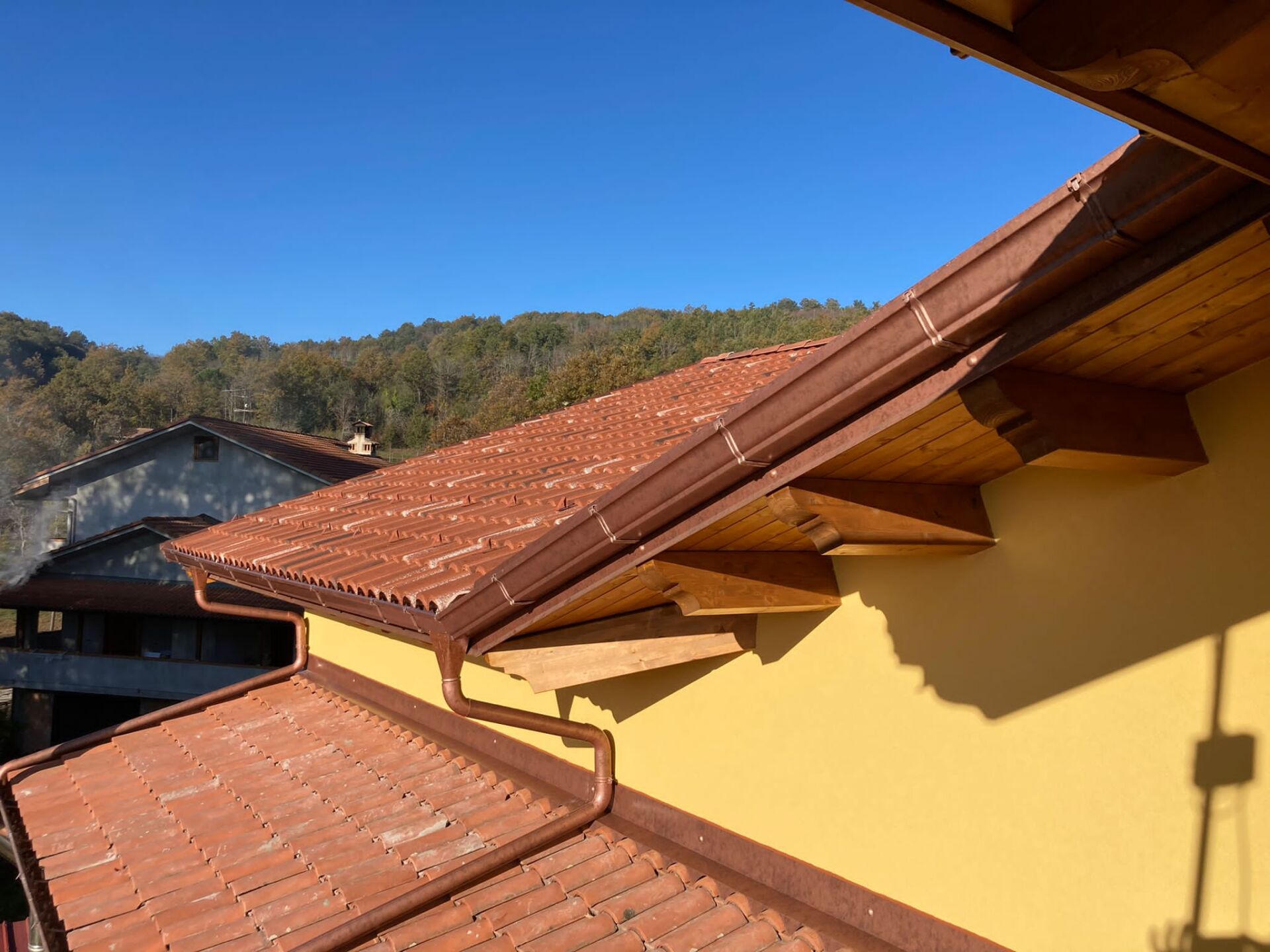 Tetto di tegole rosse con grondaie marroni e sporgenza in legno contro un muro giallo, con cielo azzurro e colline boscose sullo sfondo.