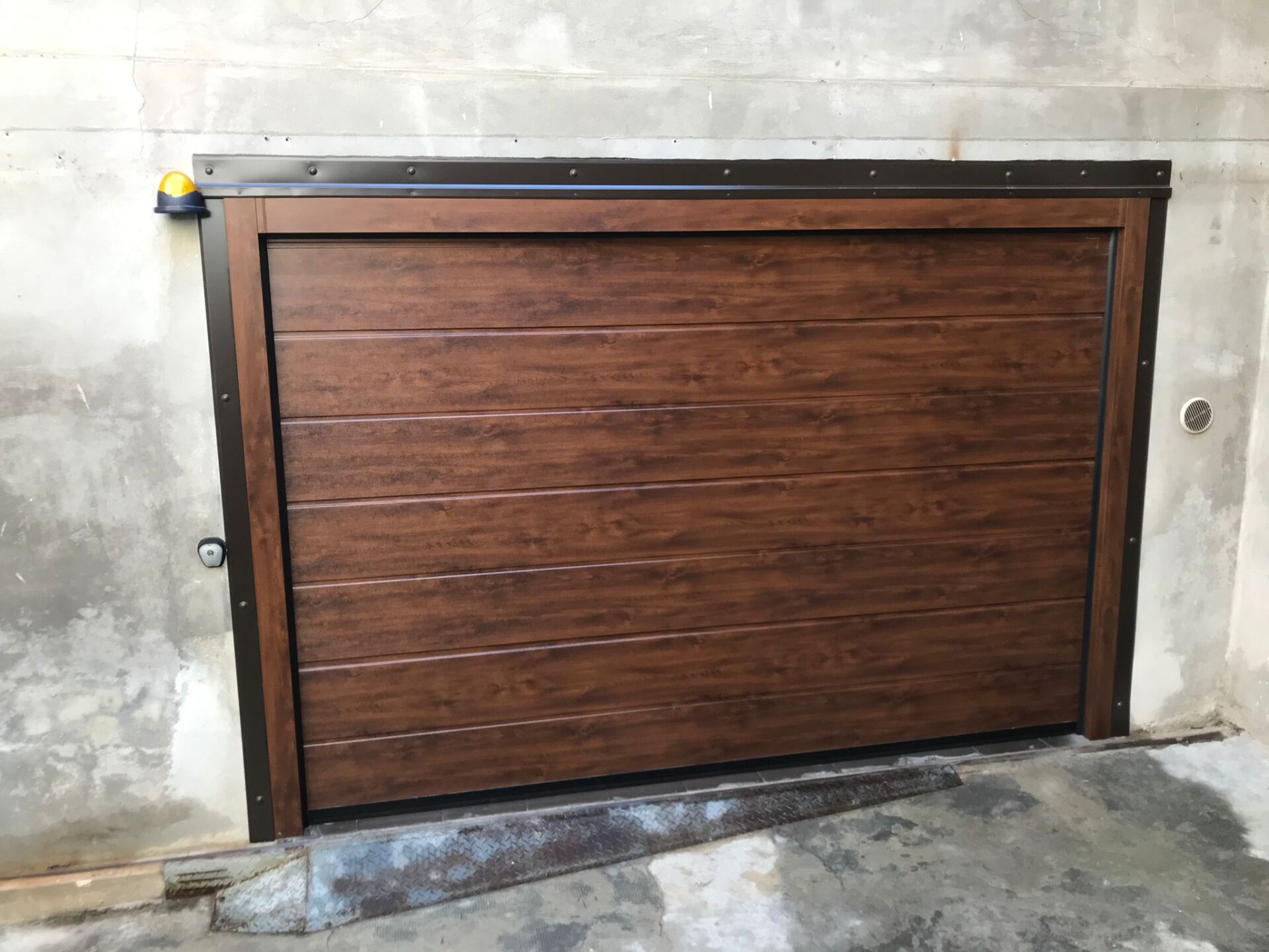 Porta da garage marrone effetto legno, incastonata in un muro di cemento, con telaio e finiture marrone scuro.