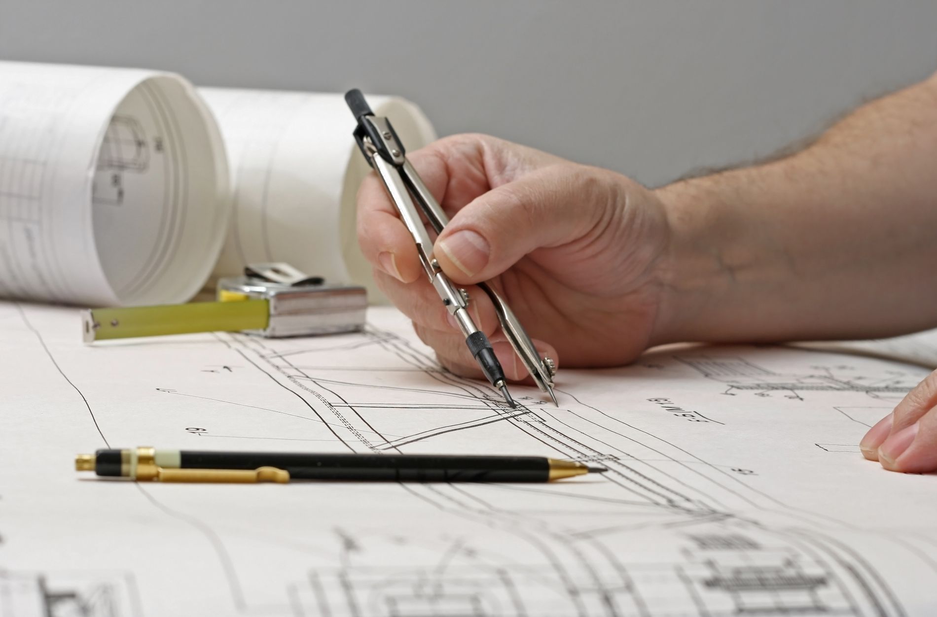 Mano che usa il compasso per disegnare su progetti architettonici, insieme a progetti arrotolati e penna.