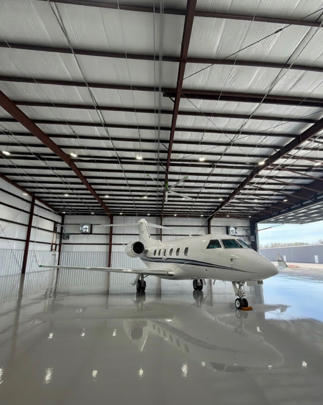 Construction site: Exterior of a gray metal building with a concrete walkway. Brown metal framework extends to the right. Cloudy sky.