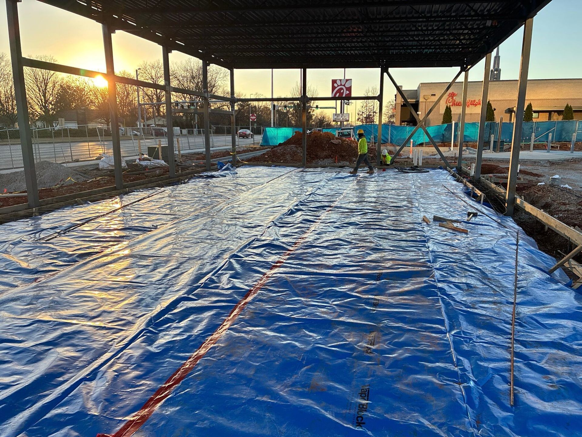 Construction site with steel frame. Blue tarp covers the ground, workers in yellow vests.