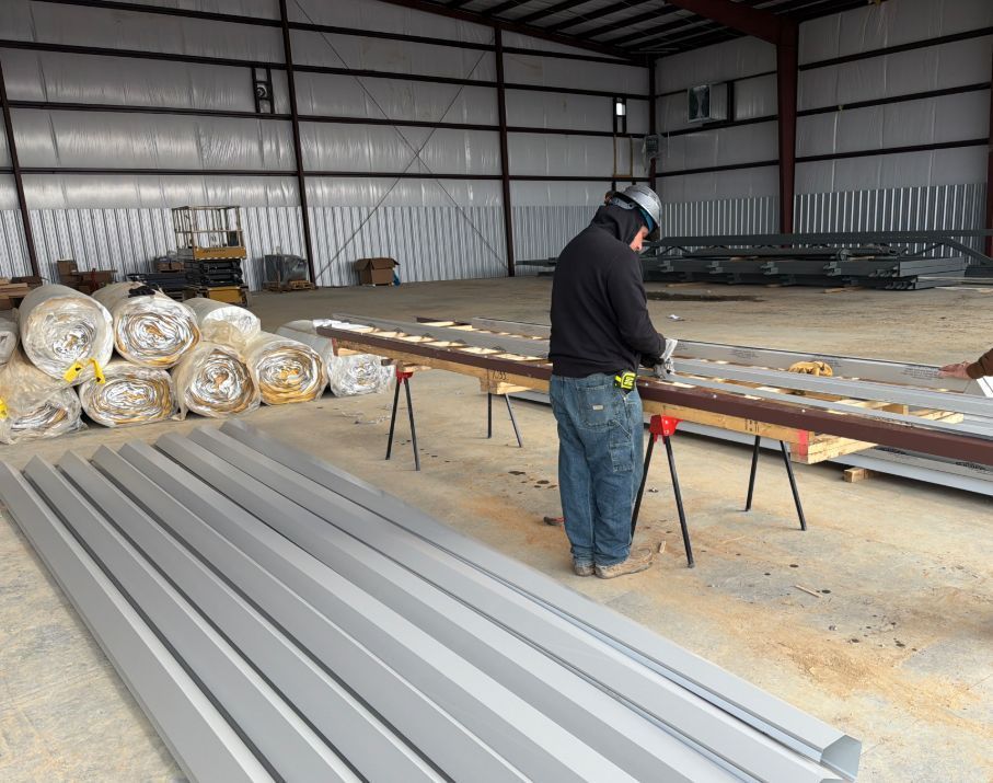 Worker cutting metal panel with saw in a large warehouse. Rolls of insulation and metal panels nearby.