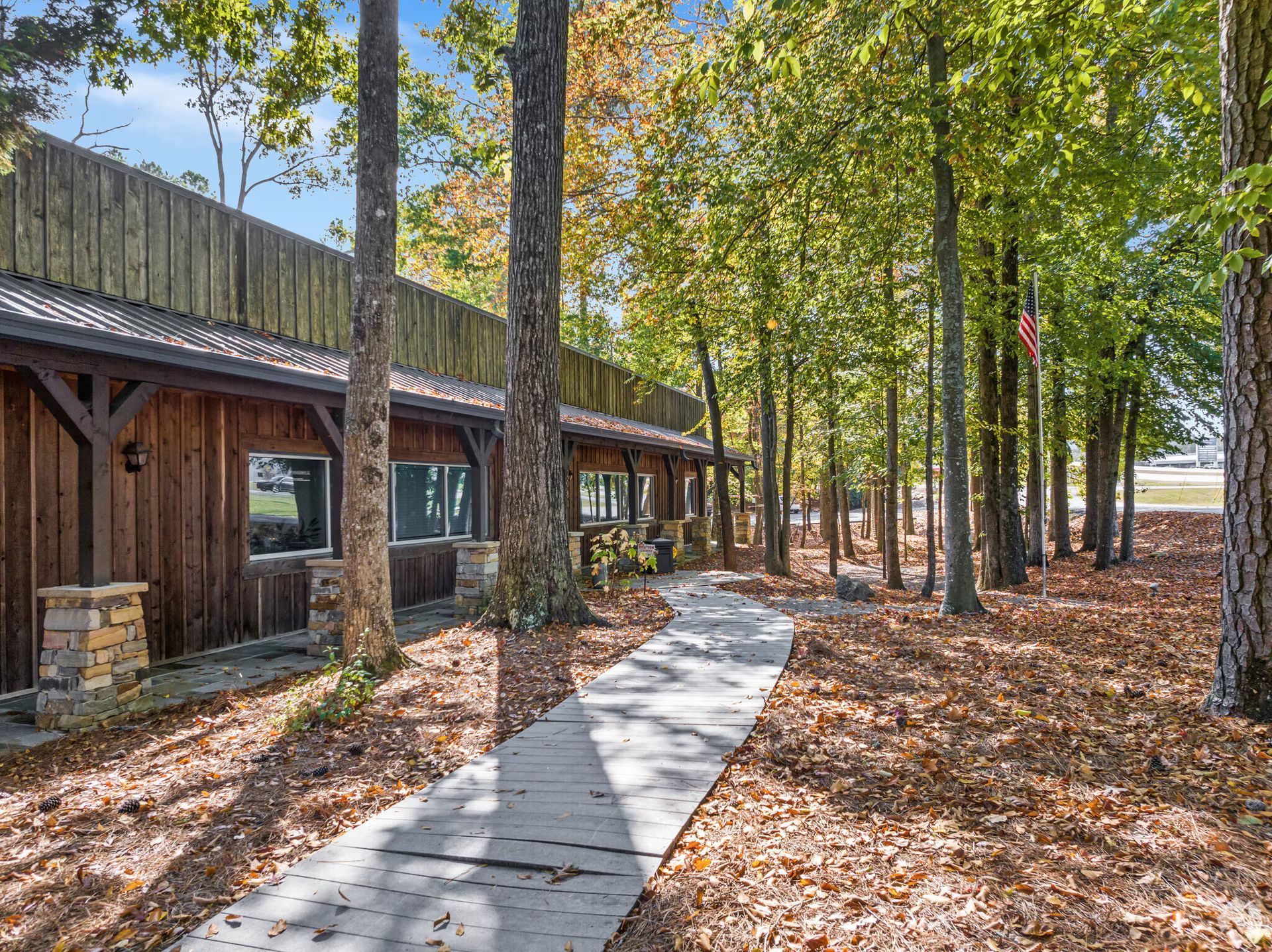 A long, rustic wooden building with stone columns sits in a forest clearing, bordered by a concrete path covered in leaves.