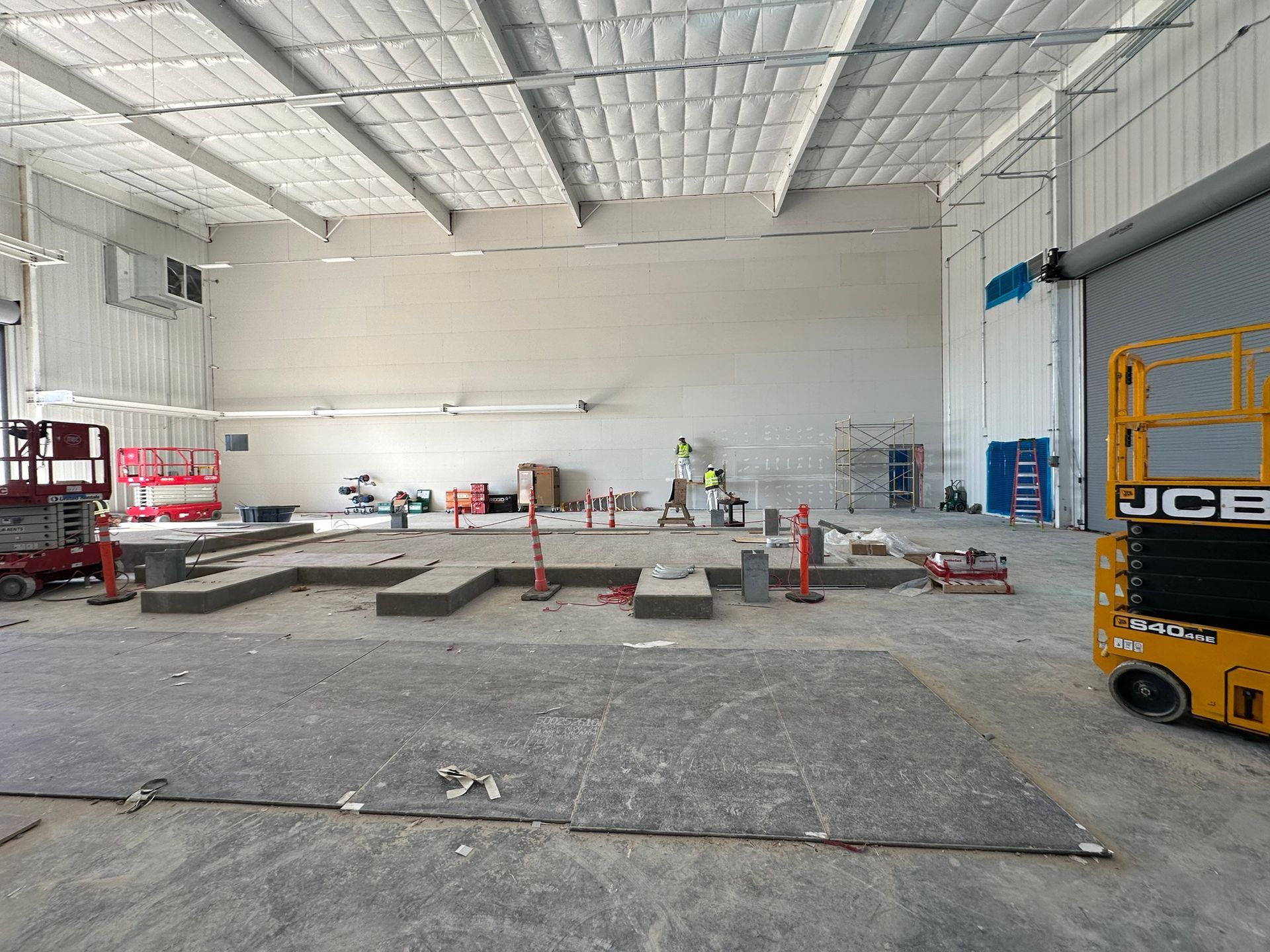 Interior of a construction site: gray concrete floor, white walls and ceiling, workers, scaffolding, and a yellow lift.