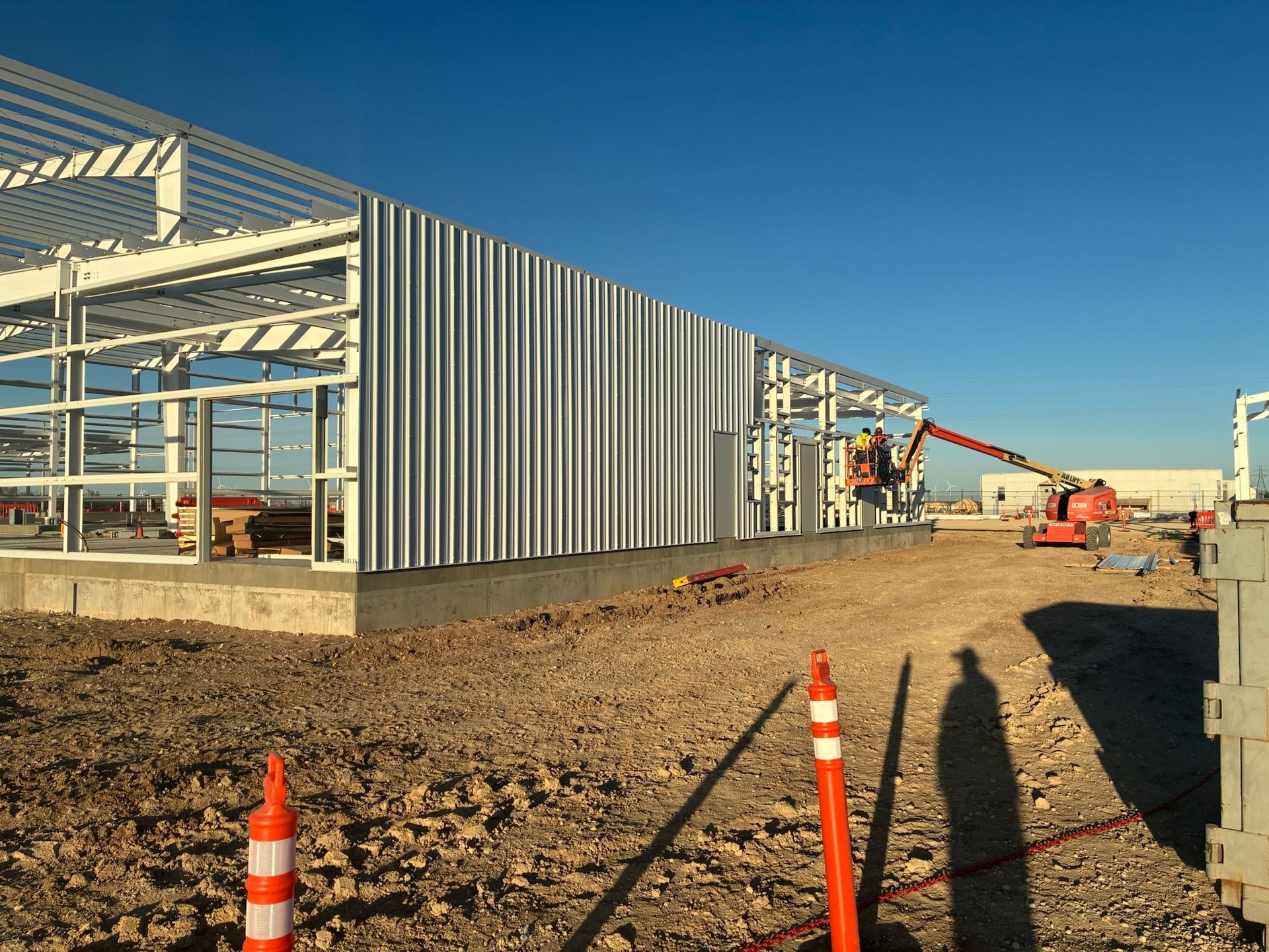 Construction site: Steel framework of a building with corrugated metal siding, workers in a lift, dirt, and blue sky.