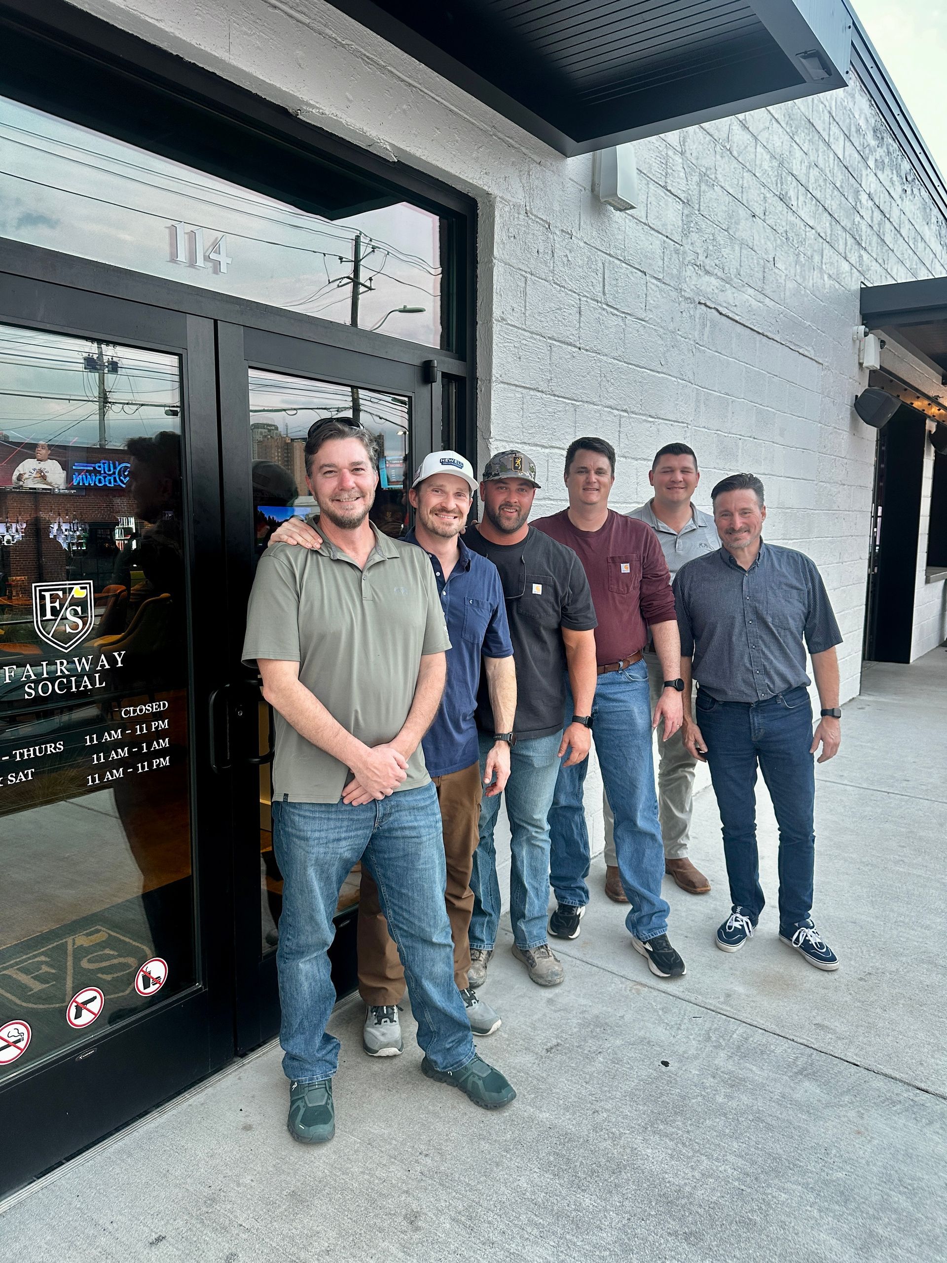 A group of six people stand together in front of a glass storefront entrance on a paved sidewalk.