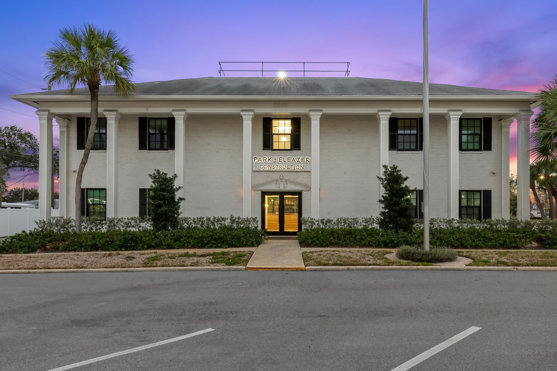 A two-story, white building with a pillared facade and dark shutters under a sunset sky, with a palm tree on the left.