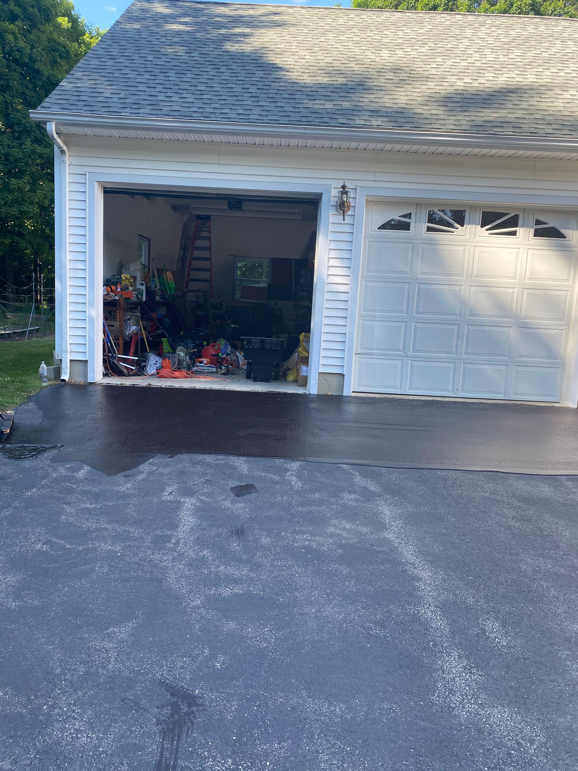 A detached two-car garage with one open bay showing storage clutter, fronted by a freshly sealed dark asphalt driveway.