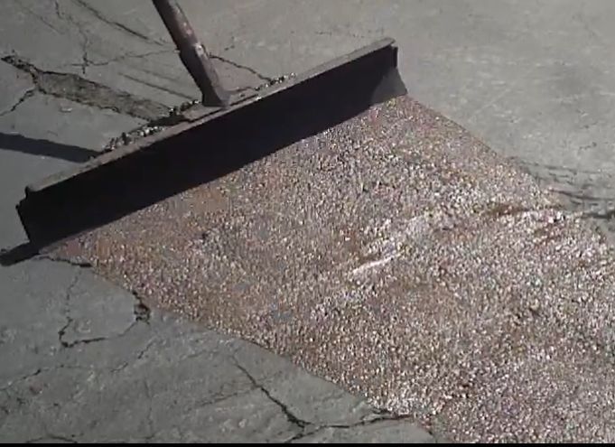 A tool spreads wet, dark gray patch material over cracks in a paved surface.