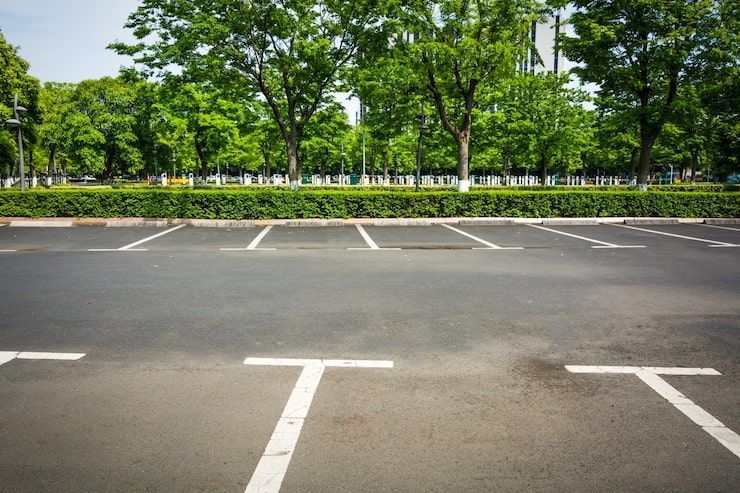 An empty parking lot with marked spaces in the foreground, bordered by a green hedge and trees in the background.