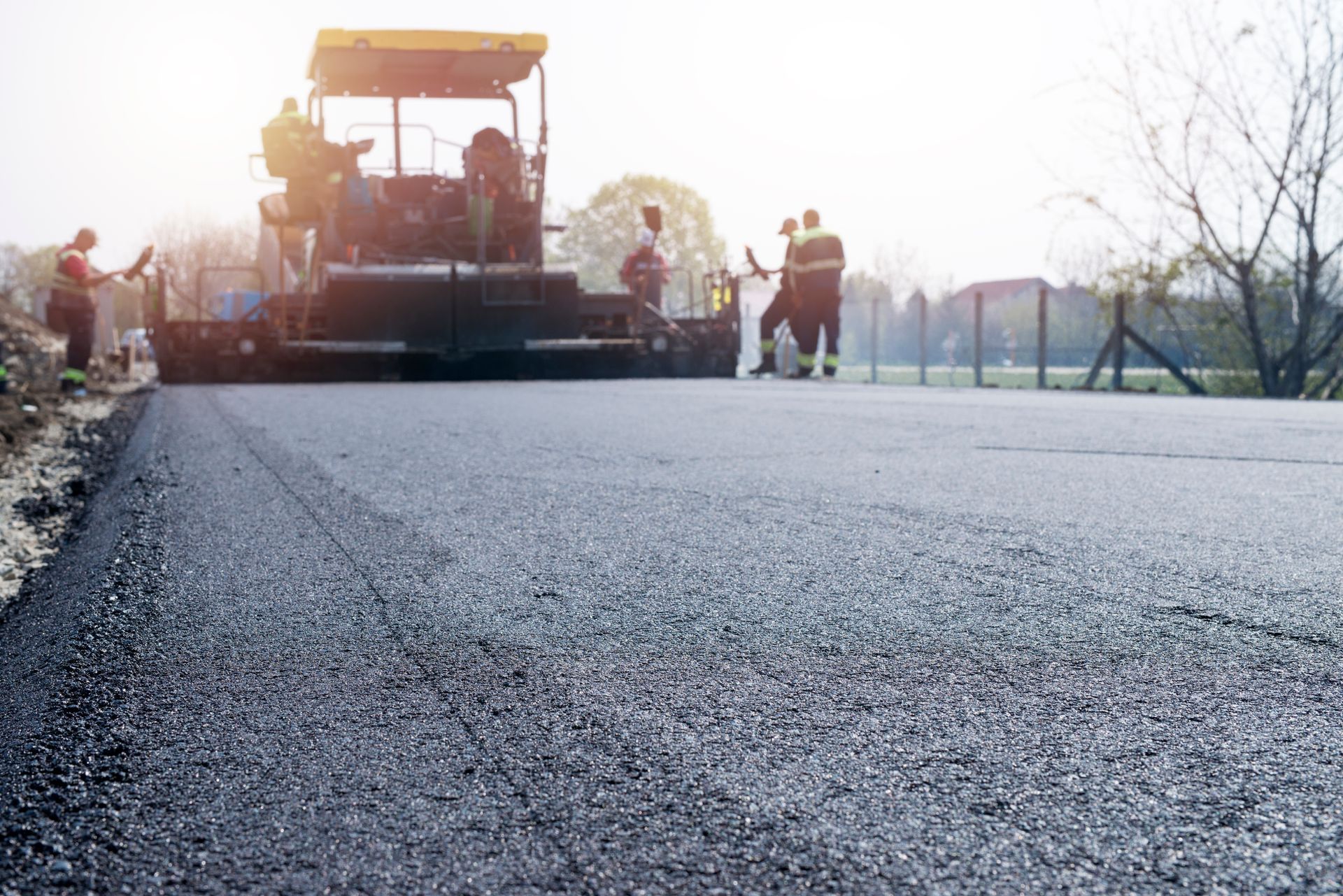Construction workers operating machinery to pave a road with fresh black asphalt under bright daylight.