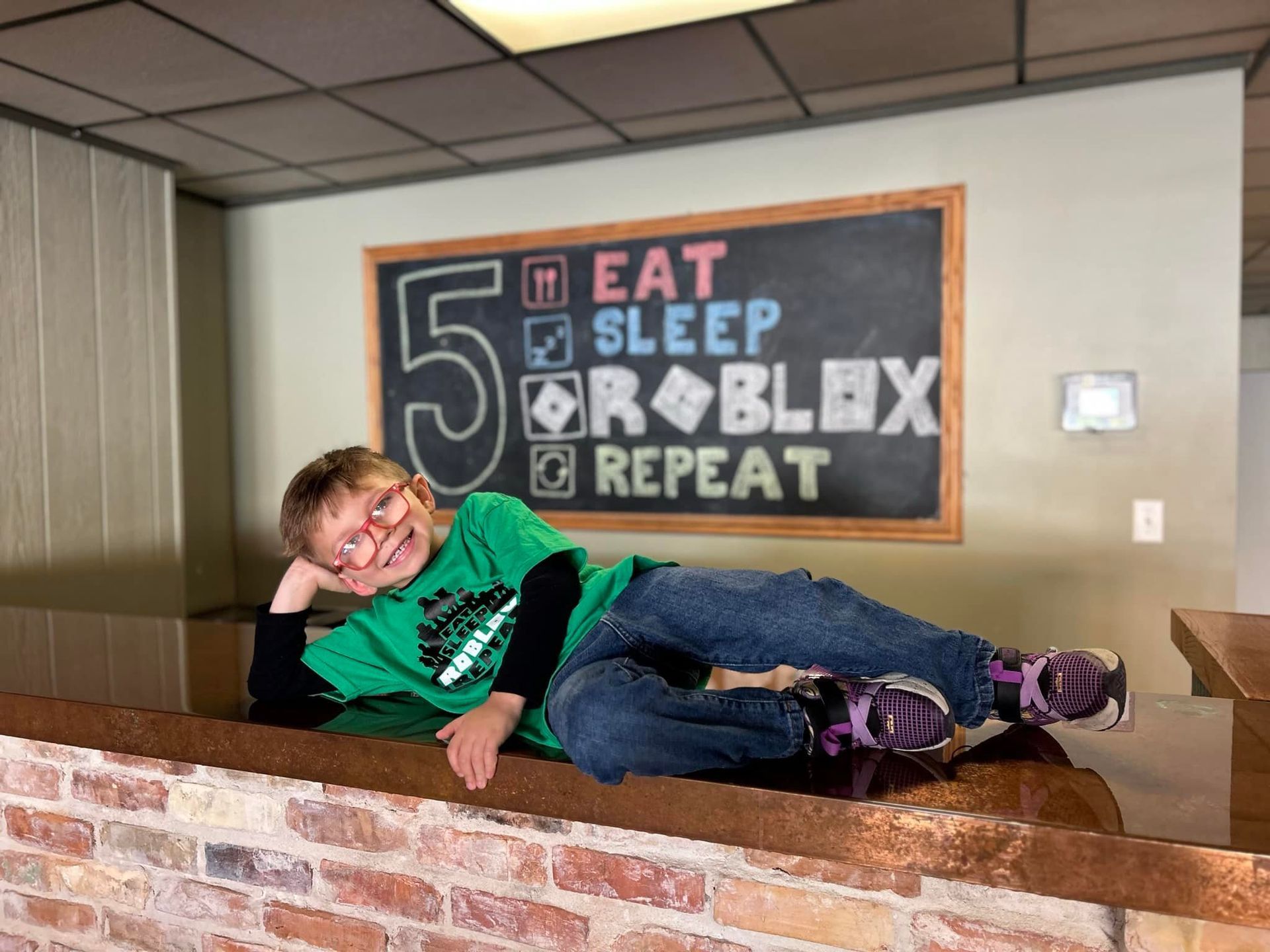 A young boy is laying on a brick wall in front of a sign that says eat sleep roblox repeat.