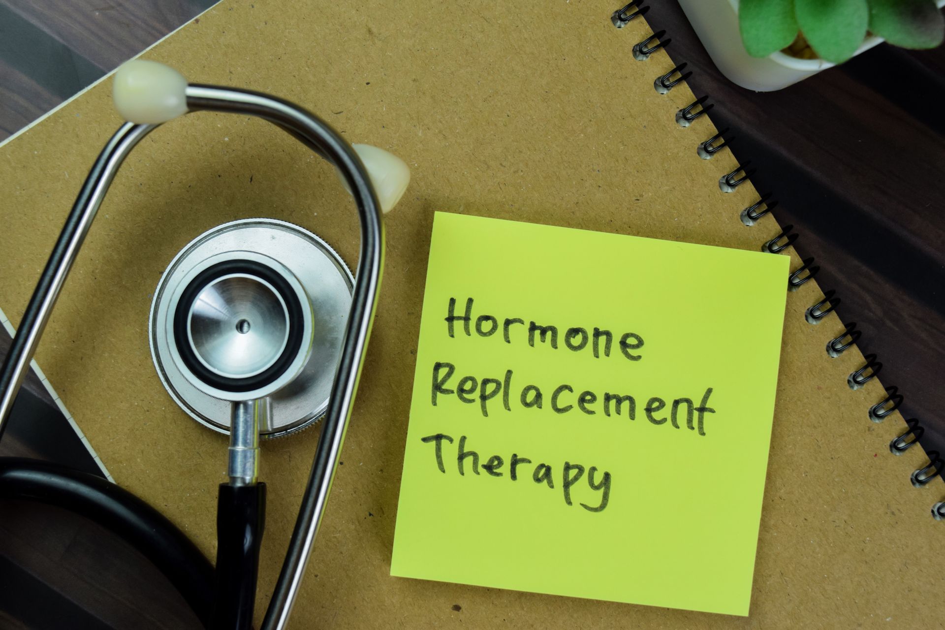 A stethoscope lies next to a lime-green sticky note on a brown notebook, with the text Hormone Replacement Therapy.