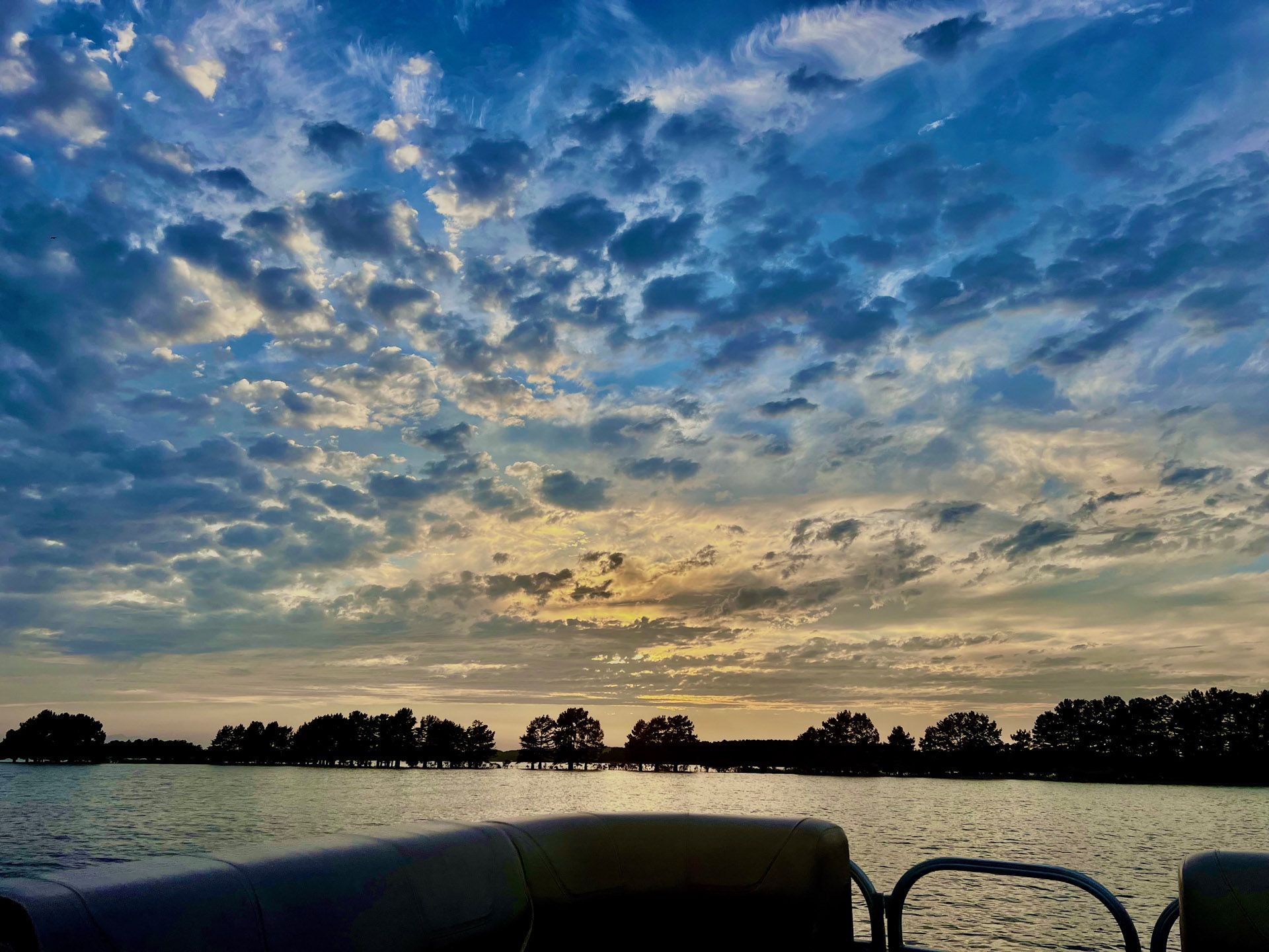 Sunset over a lake with a cloudy blue sky; trees in silhouette along the horizon; seen from a boat.