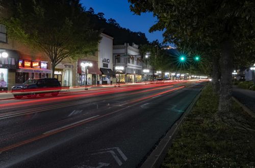 Nighttime street scene with light trails from cars. Buildings and trees line the road; a dark mountain is in the background.
