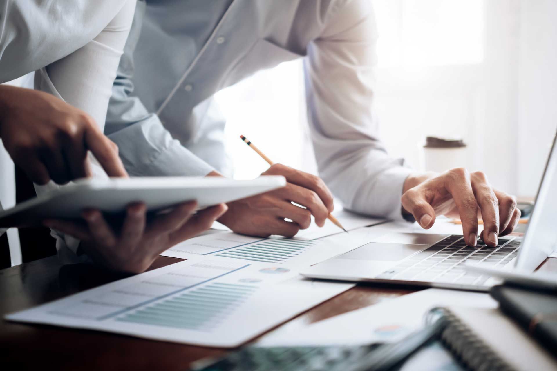 Two people reviewing financial reports at a desk, one using a laptop, the other holding a tablet.