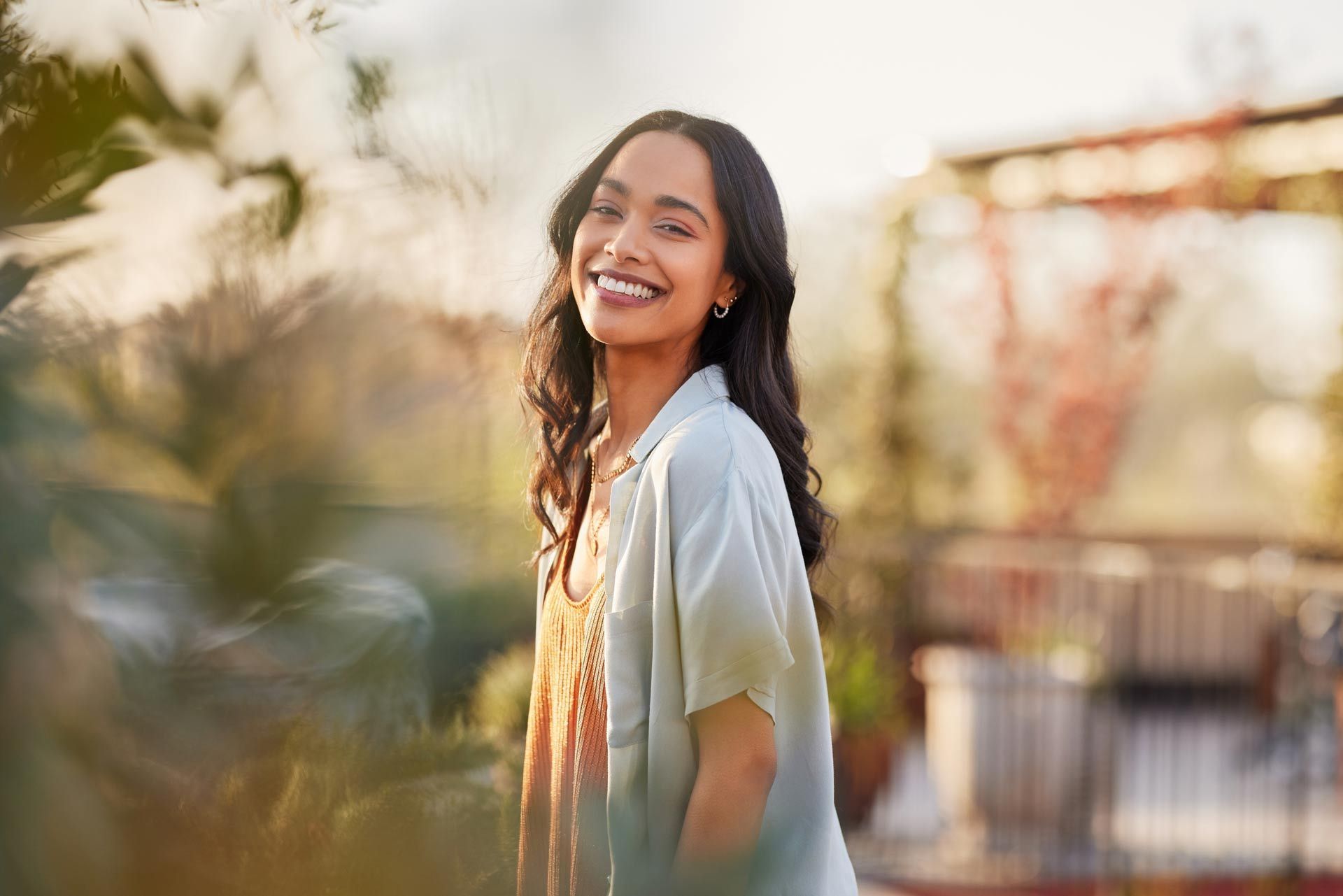 Woman smiling outdoors, wearing a light blue shirt and orange dress, with plants in the foreground.