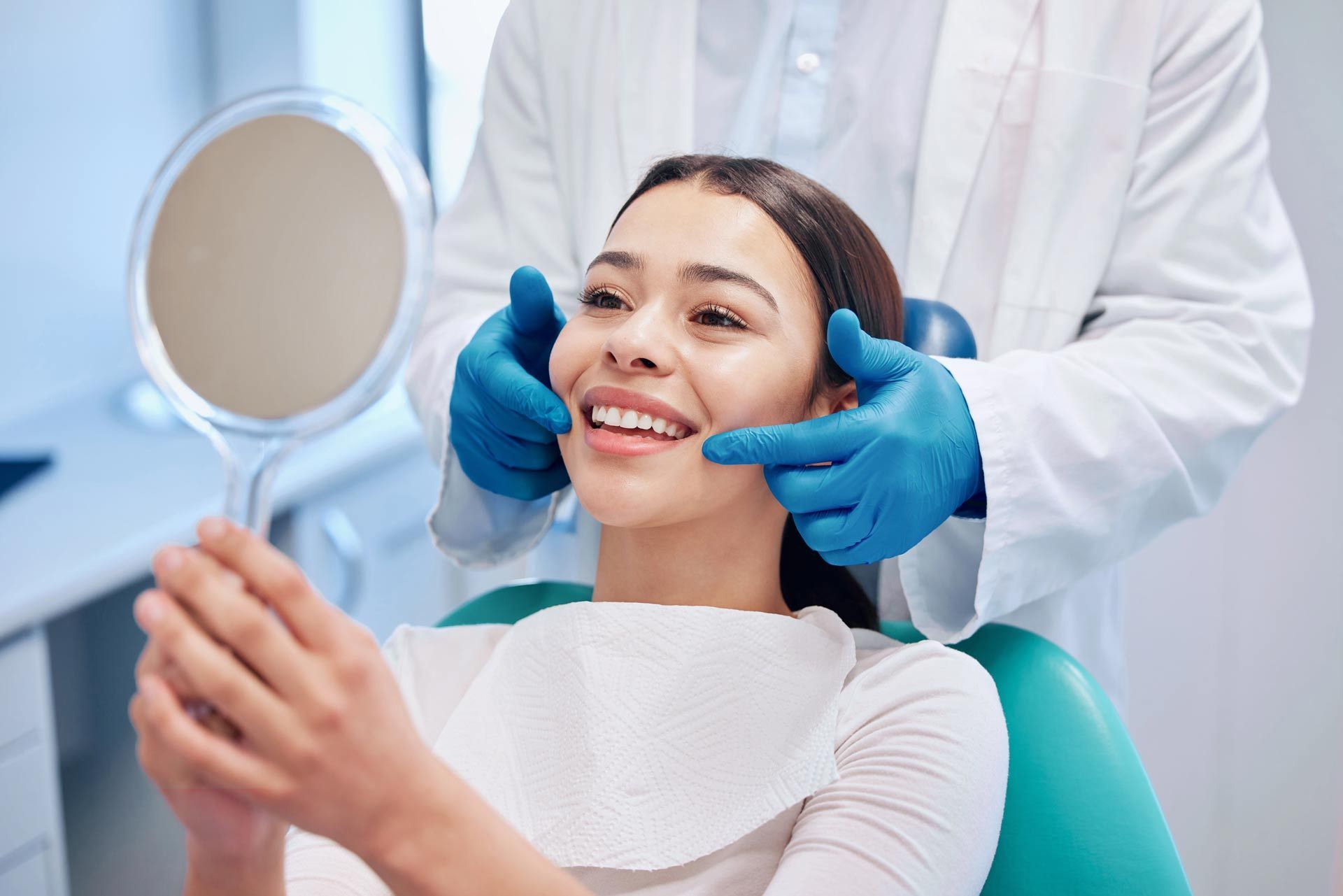 A dentist examining a smiling woman's teeth with a mirror. White coat, blue gloves, dental office.
