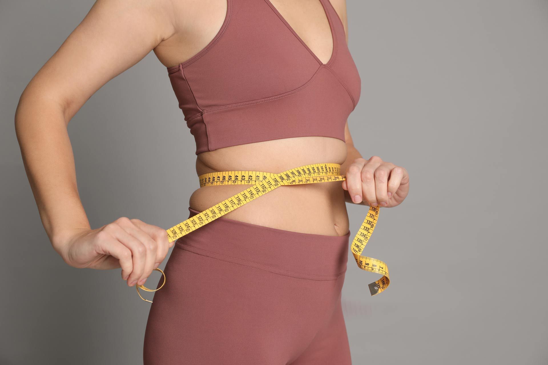 Woman in pink athletic wear measuring waist with yellow tape against a gray background.