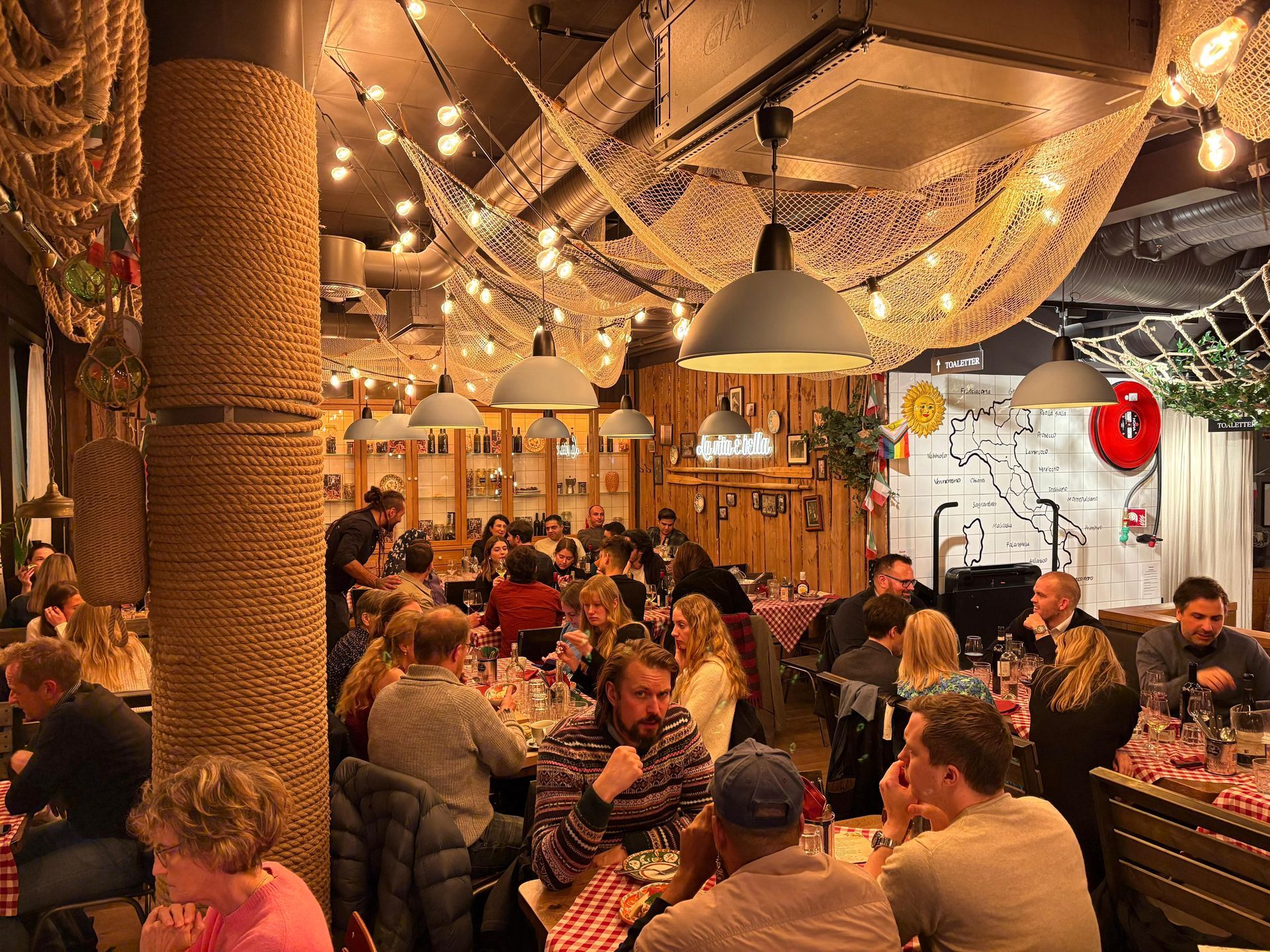 Restaurant interior with many people dining; nautical decor, rope-wrapped columns, overhead string lights.