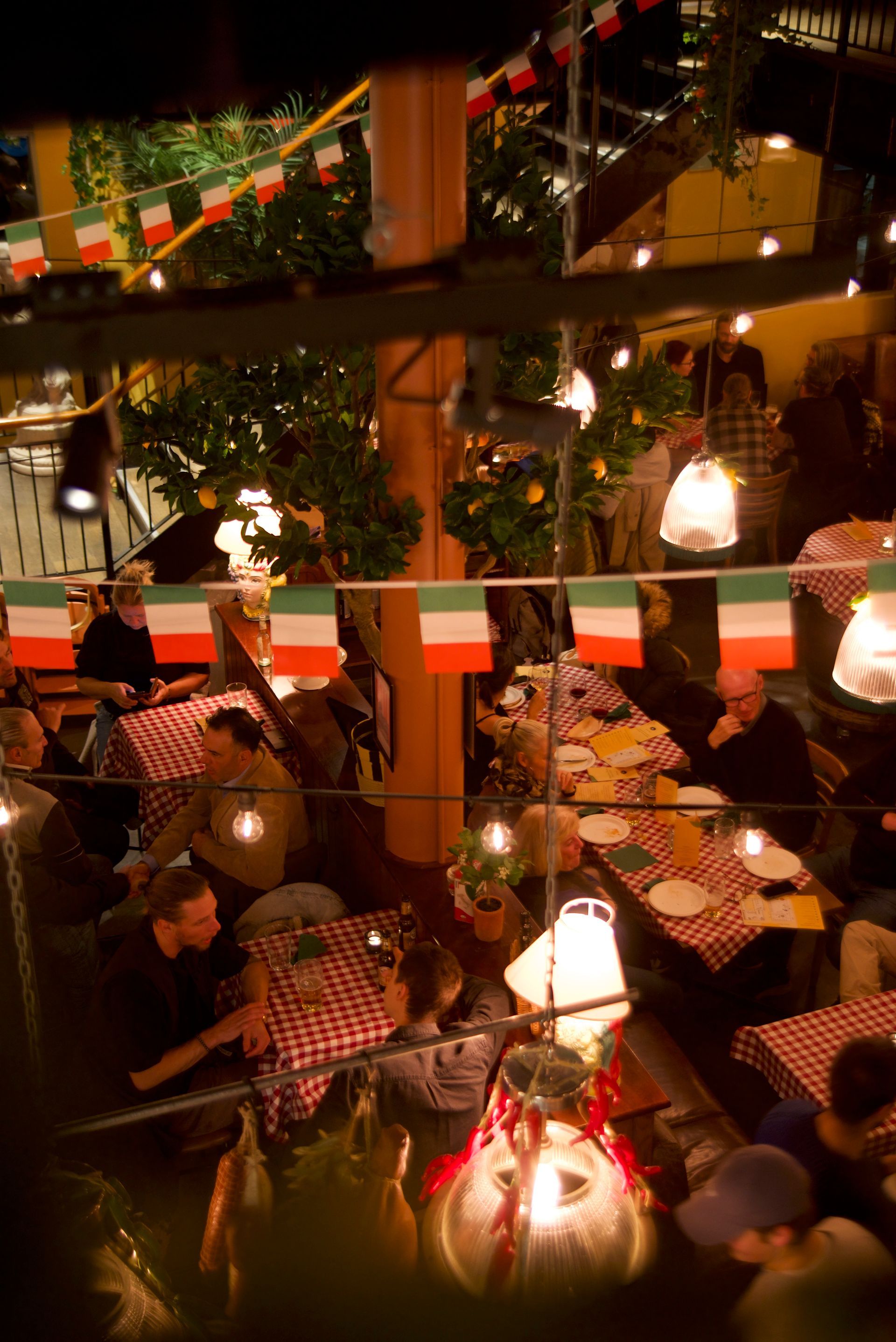 Cozy restaurant interior with patrons at checkered tablecloth tables under string lights and a neon sign.