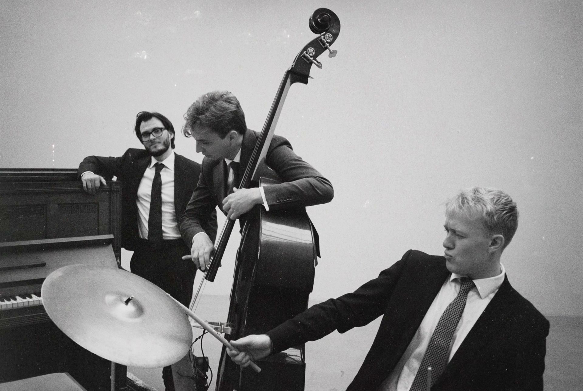 A jazz trio: pianist, bassist, and drummer in suits performing, with instruments. Black and white photo.