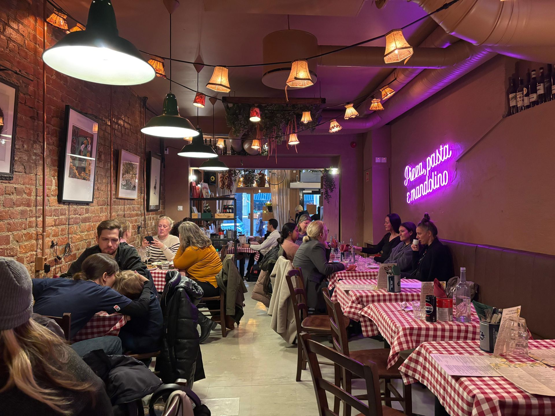 Cozy restaurant interior with patrons at checkered tablecloth tables under string lights and a neon sign.