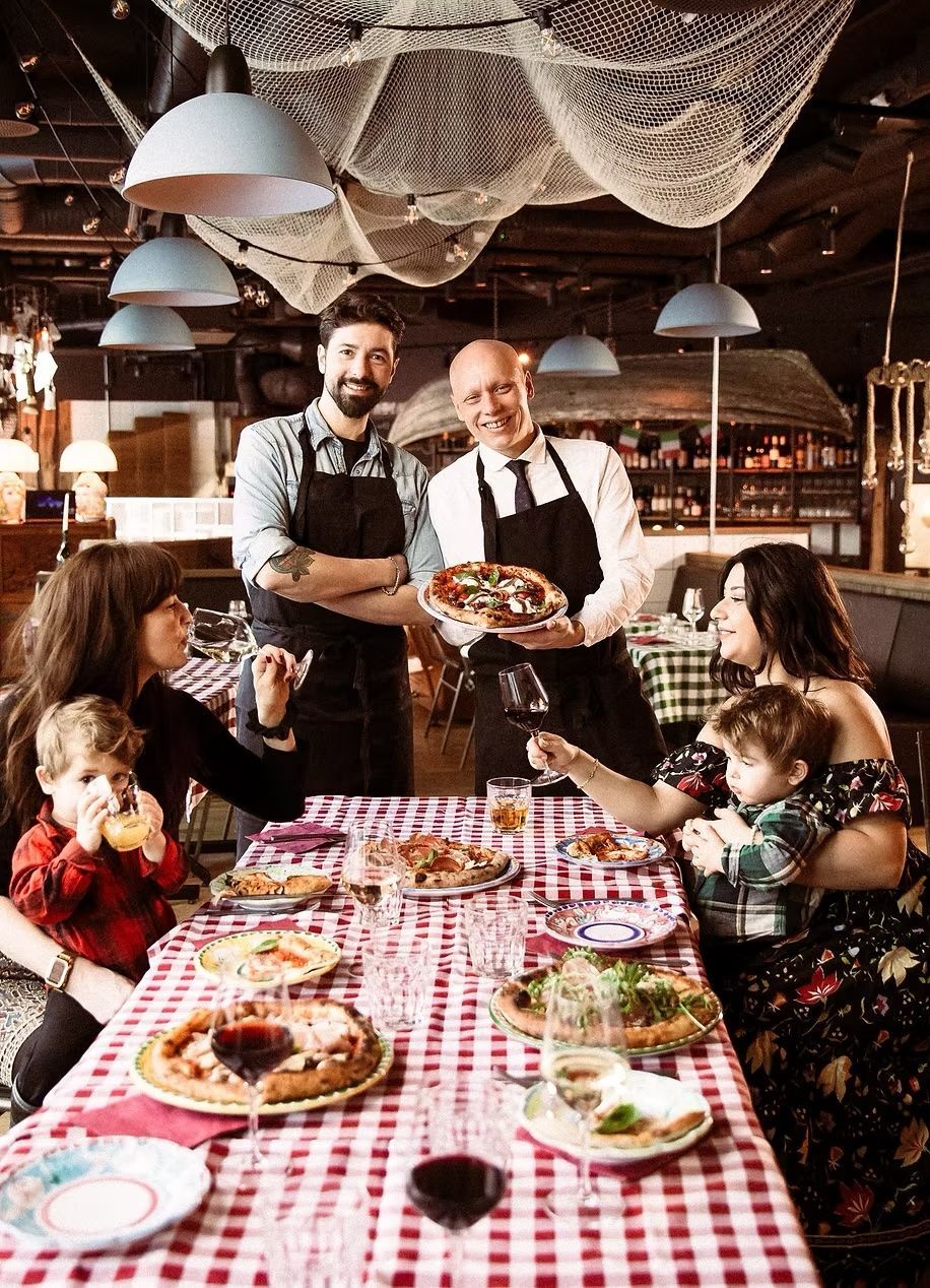 Family dining at a restaurant. Two chefs holding pizza. Red and white checkered tablecloth.