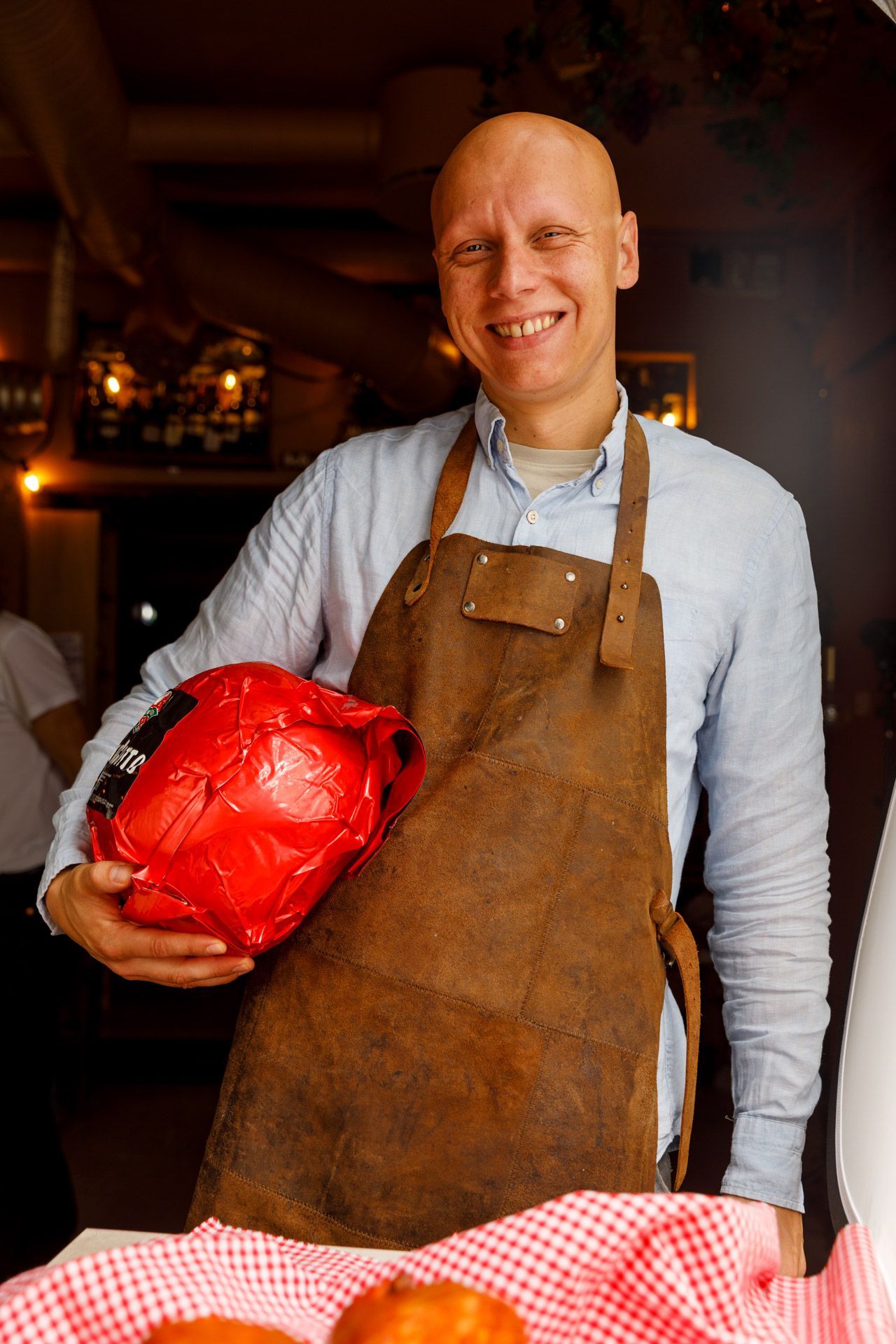 Bald man in brown apron holding a red-wrapped ham; smiling in a restaurant setting.