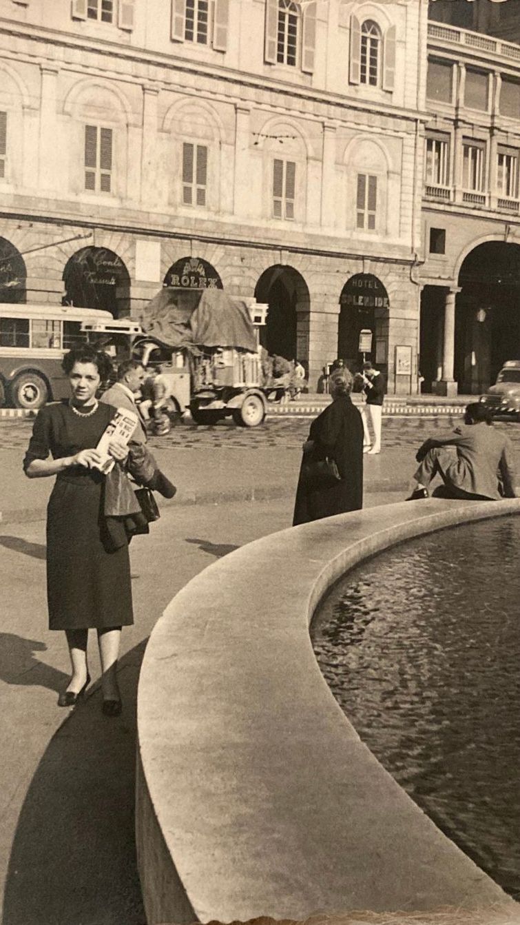 Woman holding a child stands near a fountain in a European square. A car and people are in the background.