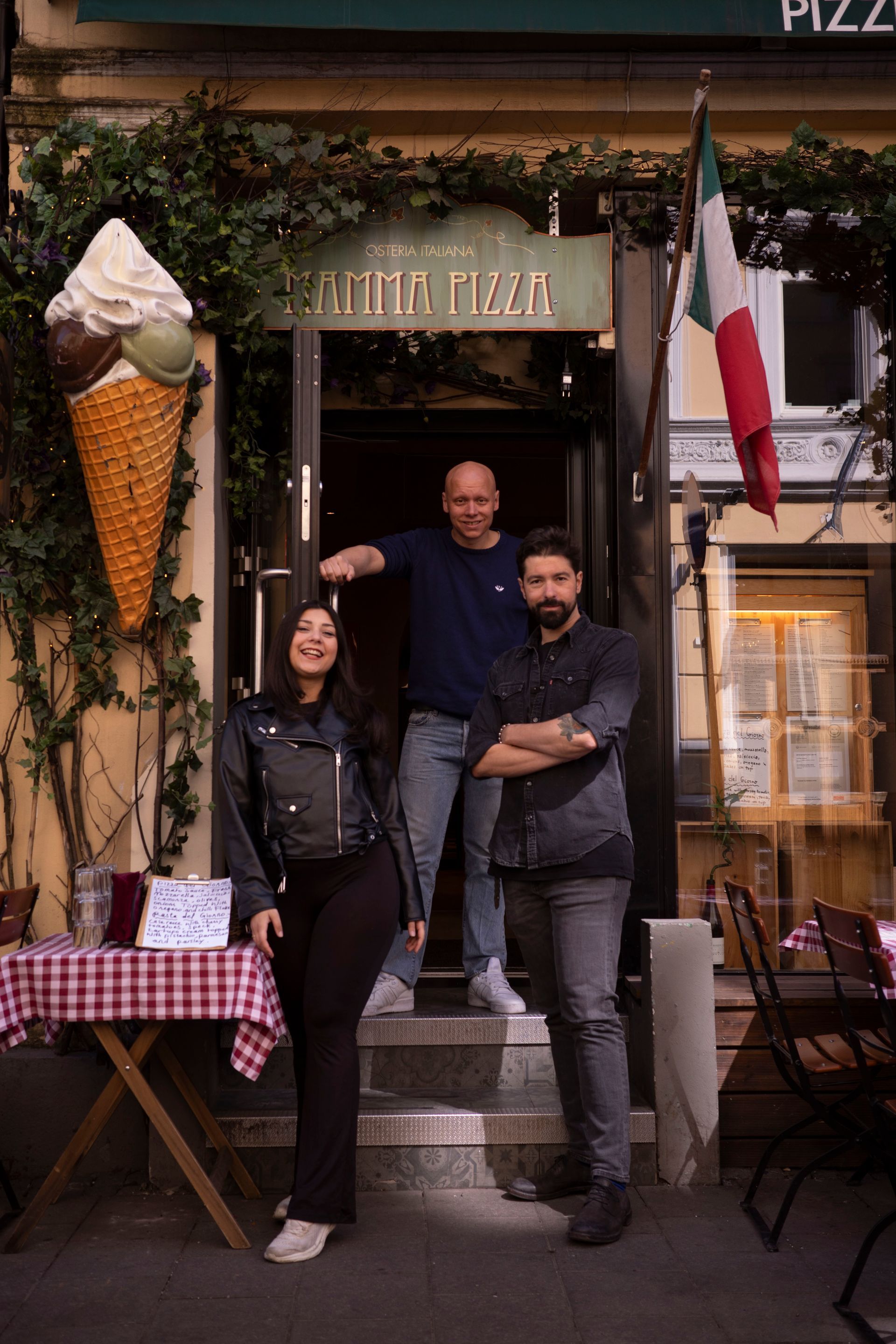 Three people outside Italian pizzeria. Woman, man and man. Building has giant ice cream cone and Italian flag.