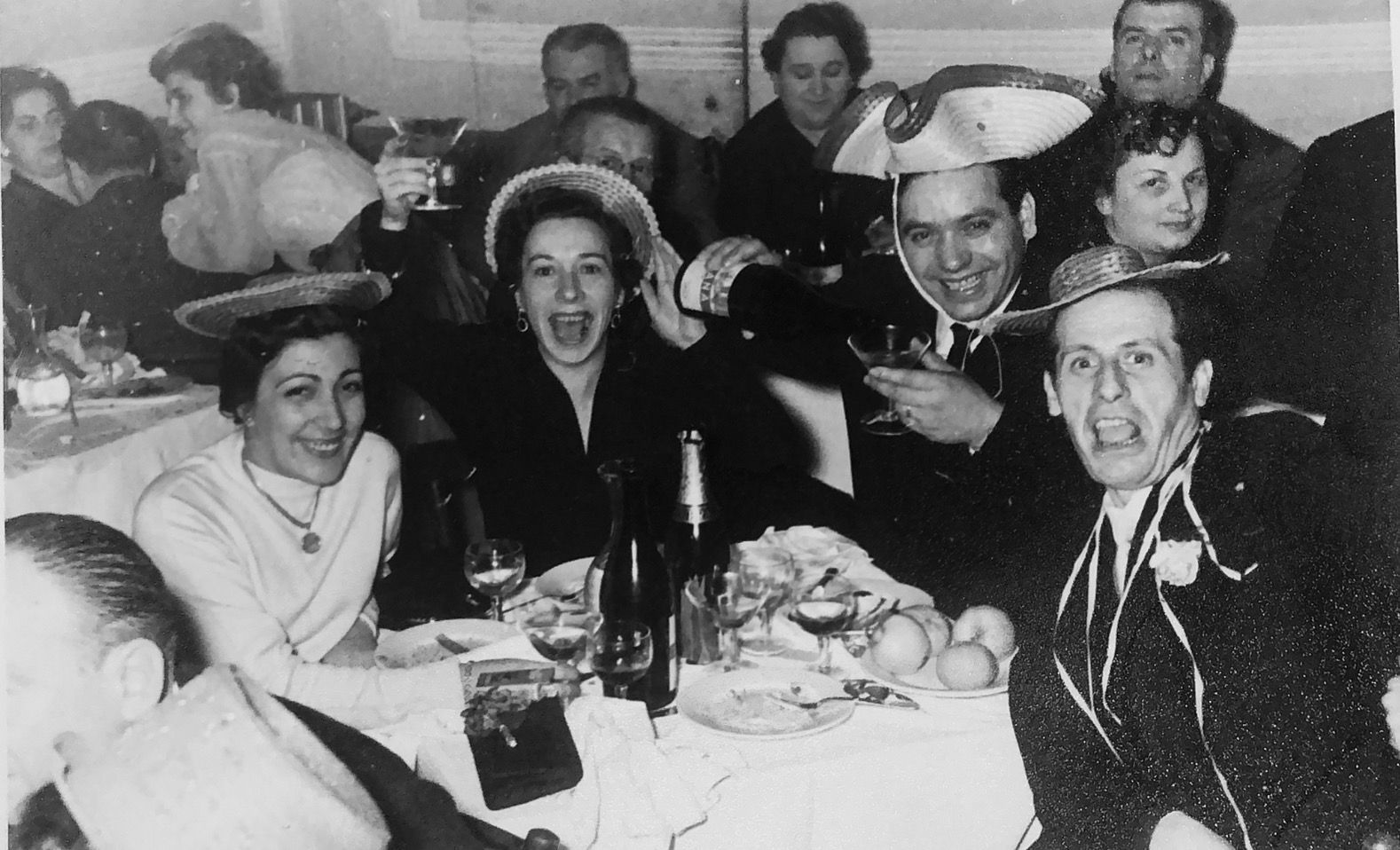 People celebrating at a table with drinks, some wearing hats, happy expressions, in a dimly lit room.