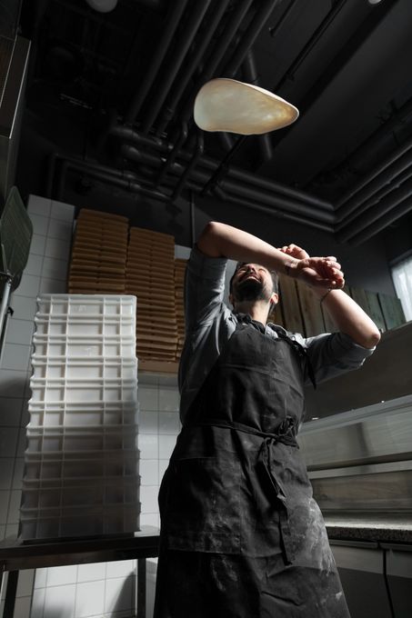 Chef tossing pizza dough in the air, dark kitchen setting.